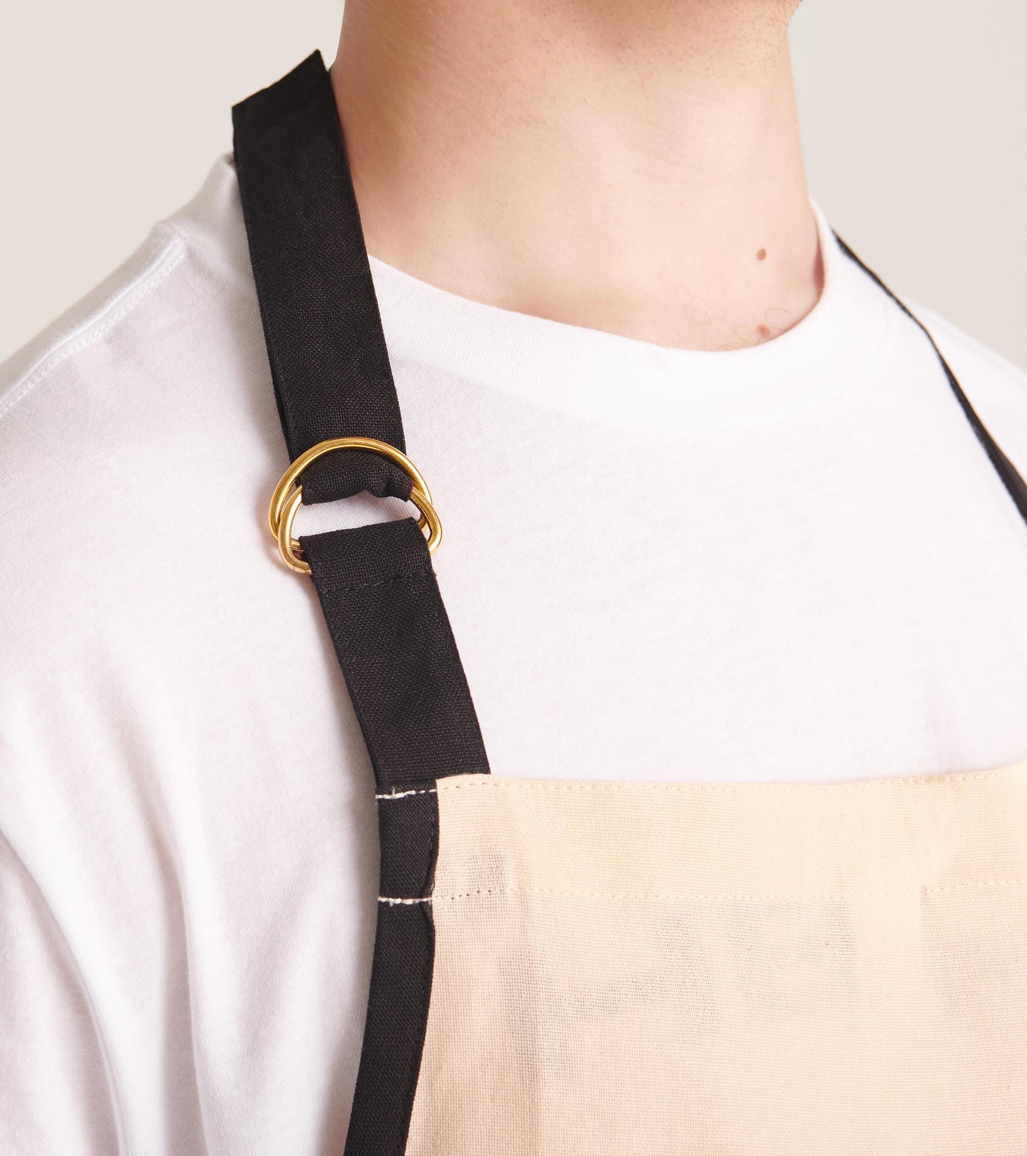 Beige apron with black straps and a gold ring detail, designed for cooking and kitchen use.