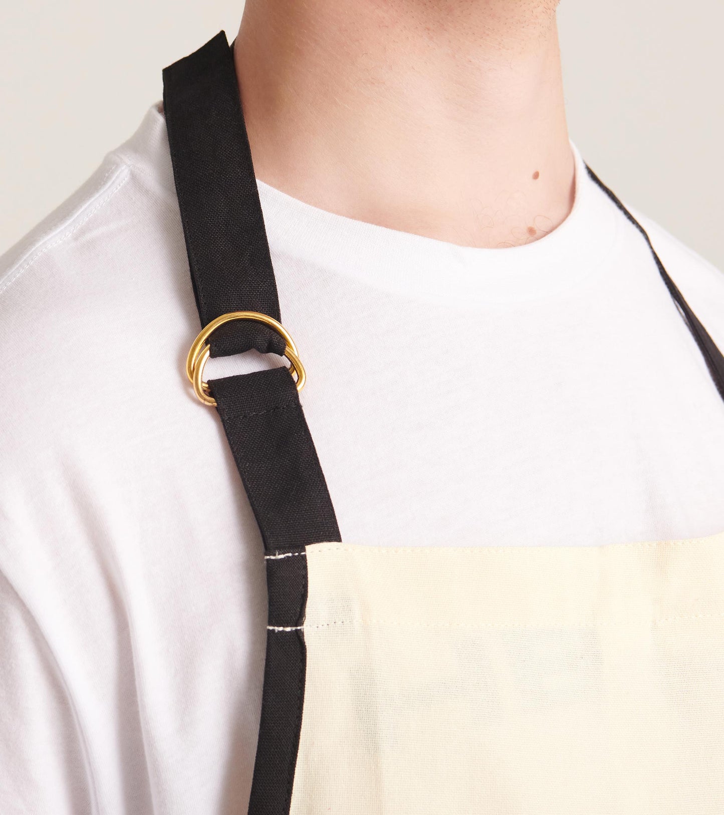 Beige apron with black straps and a gold ring detail, designed for cooking or crafting.