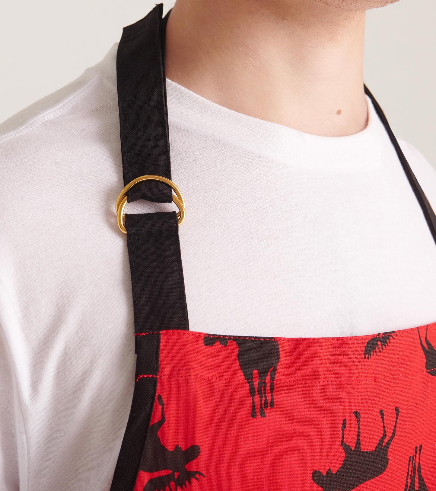 Red apron featuring black moose silhouettes, adjustable neck strap with a gold ring, worn over a white shirt.