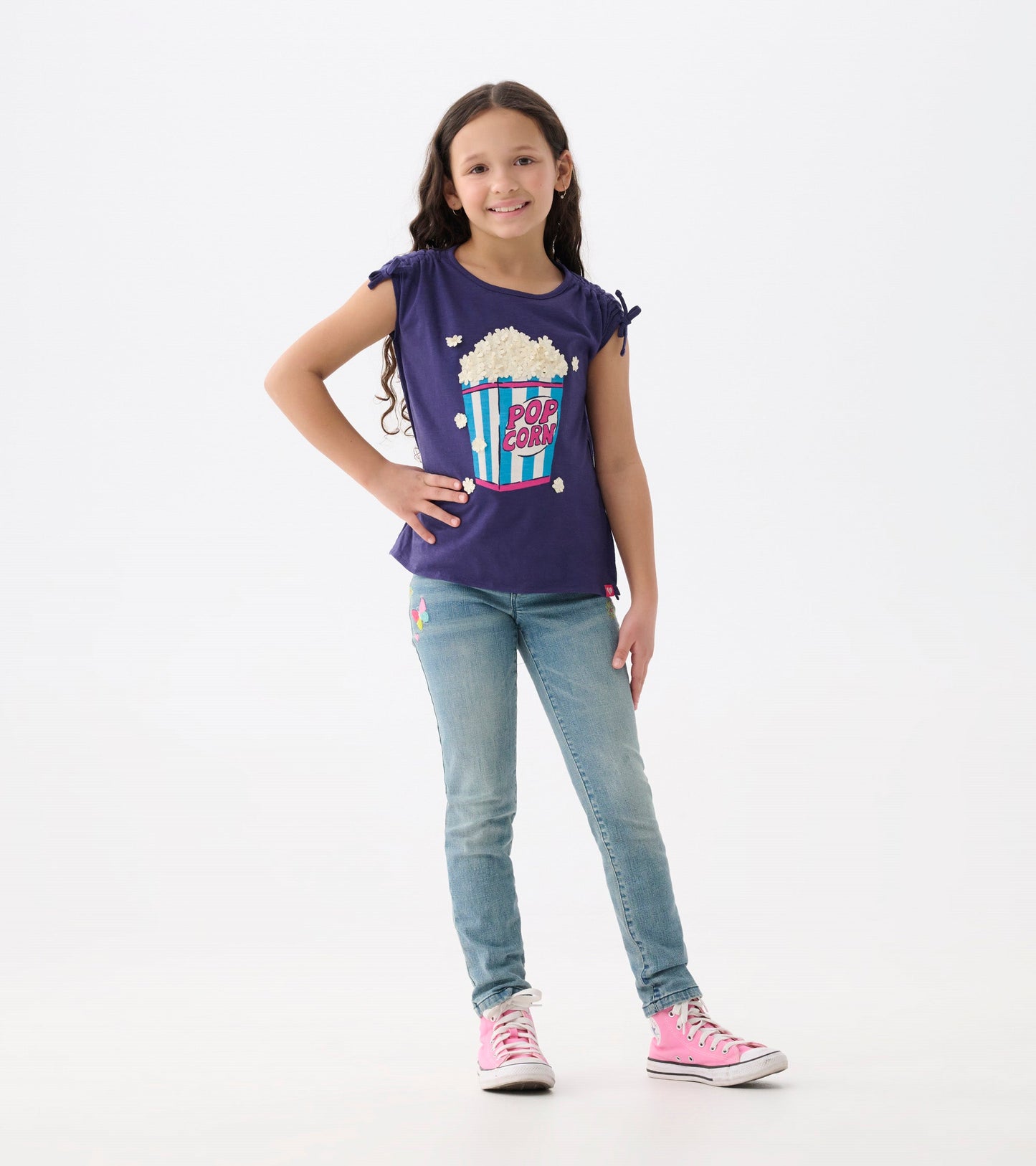 Light blue stretch jeans with colorful patches, fitted style, worn with a purple popcorn graphic tee and pink sneakers.