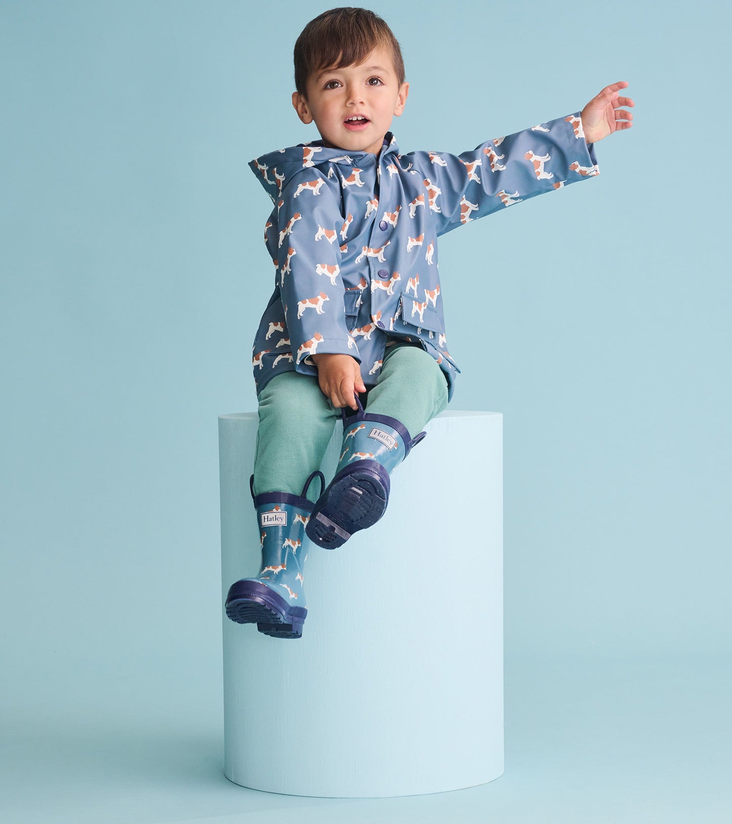 Blue rain jacket with French bulldog print, worn by a boy sitting on a pedestal, paired with green pants and blue boots.