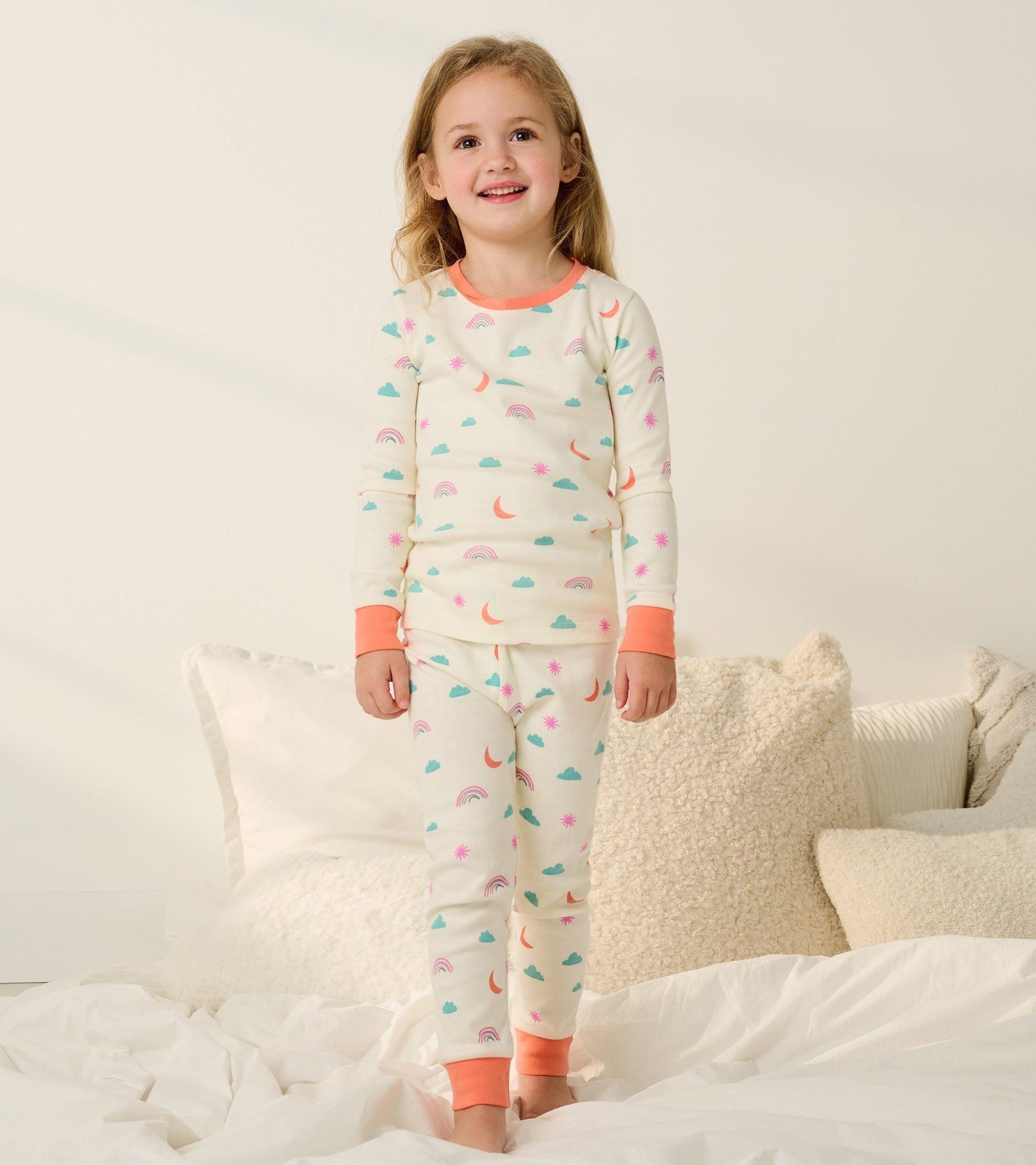 Organic cotton pajama set for girls featuring a cream base with colorful clouds, rainbows, and suns, accented with coral cuf…