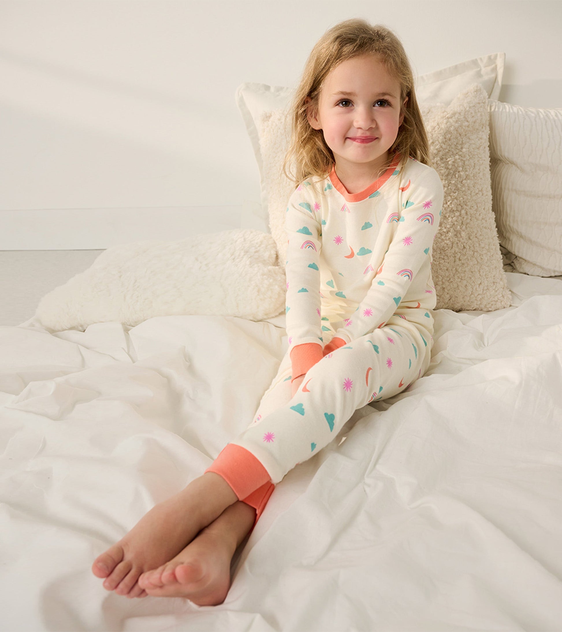 Cream-colored organic cotton pajama set with colorful cloud, rainbow, and star patterns, featuring coral cuffs and neckline.
