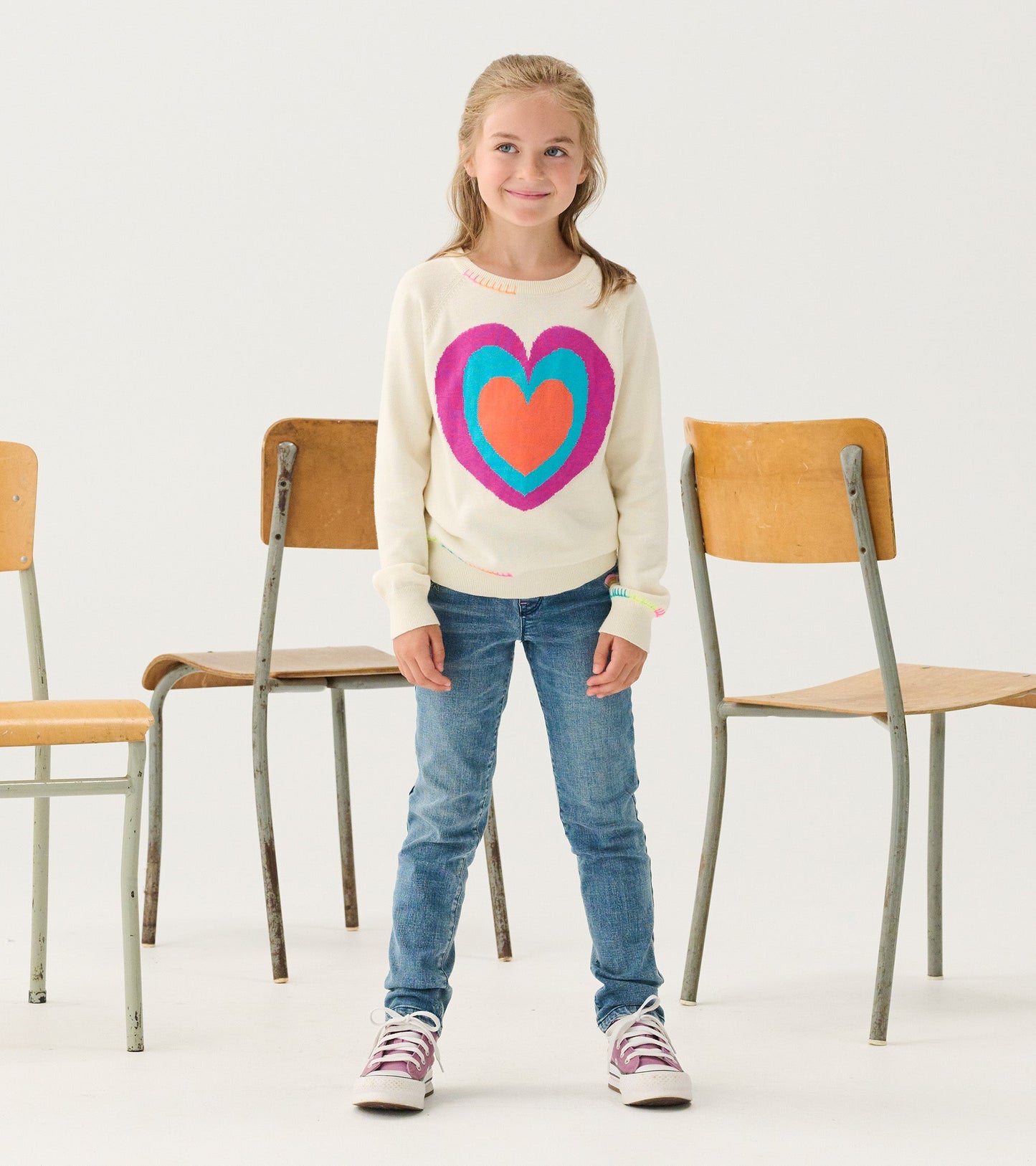 Cream sweater featuring a large layered heart design in pink and blue, paired with blue jeans and sneakers.