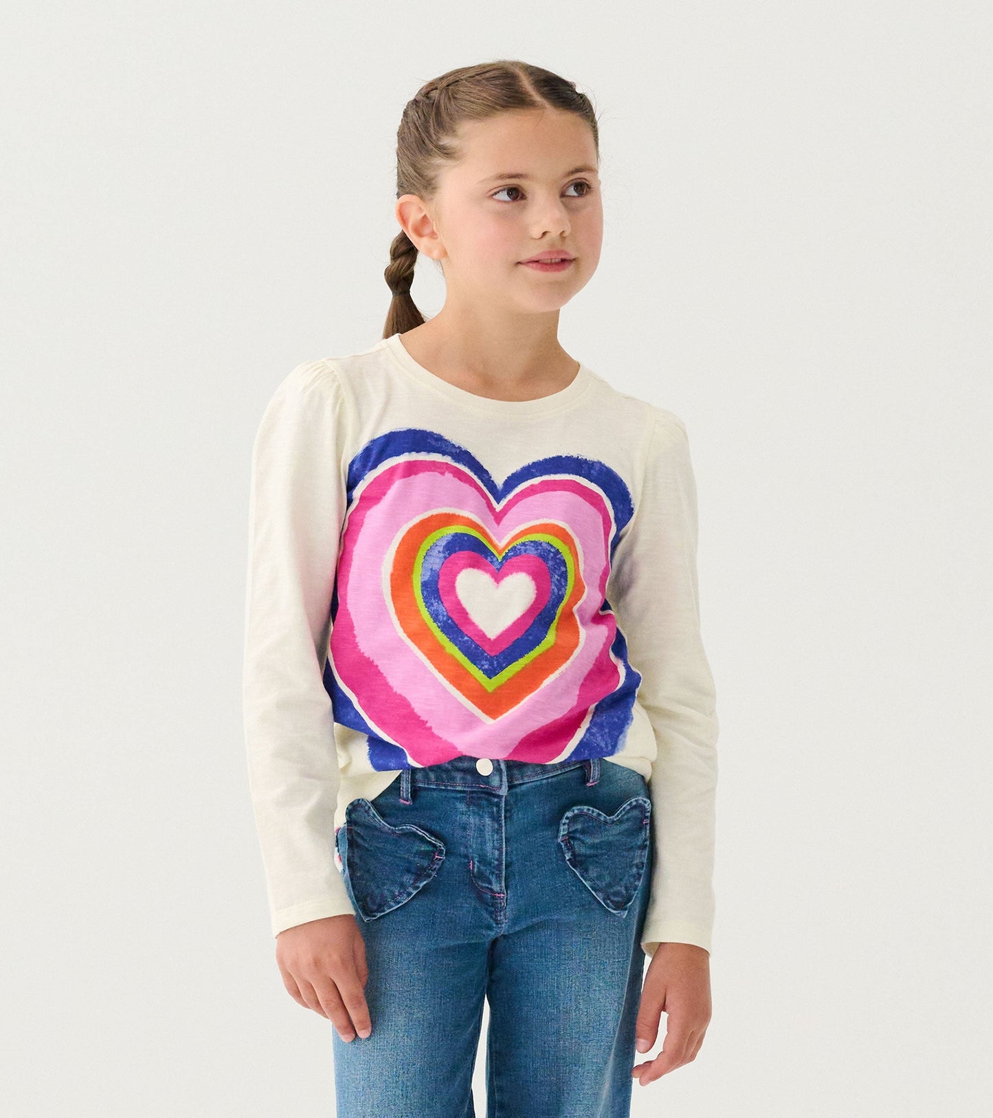 Long sleeve cream tee featuring a colorful layered heart graphic, paired with blue jeans with heart-shaped pockets.
