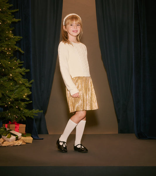Cream sweater with a textured pattern paired with a shiny gold pleated skirt, worn with white socks and black shoes.