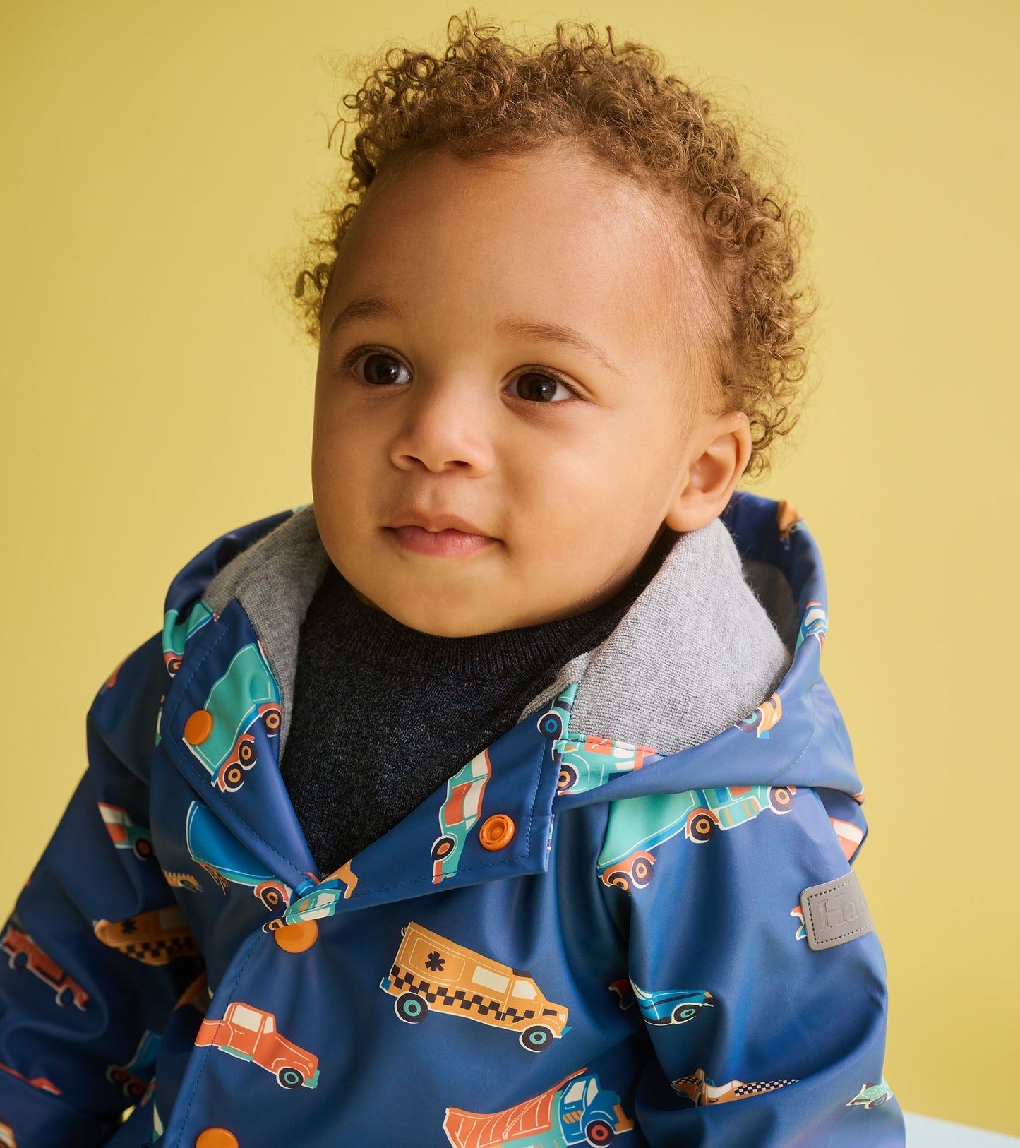 Blue rain jacket featuring colorful car patterns, with a gray hood and orange buttons, suitable for baby and toddler boys.