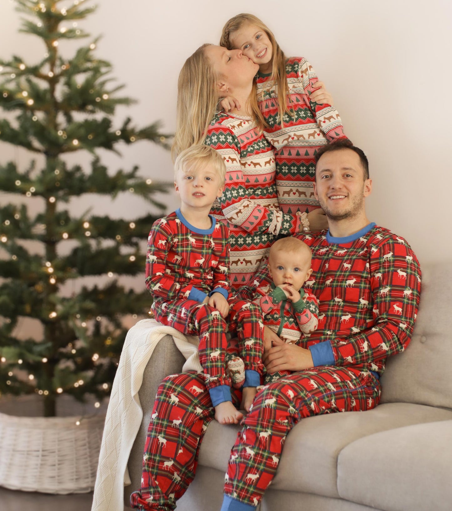 Red plaid pajama set featuring festive patterns, worn by a family of five, sitting on a couch with a Christmas tree in the b…