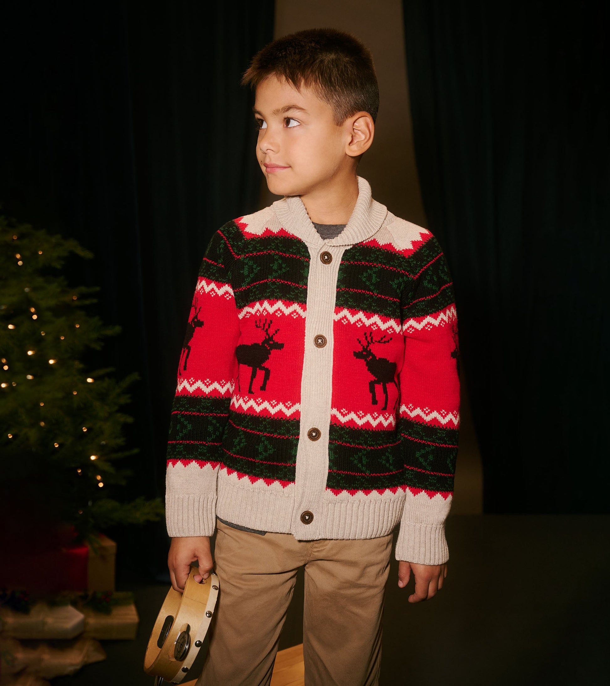 Red and green shawl collar cardigan with reindeer pattern, featuring buttons and ribbed cuffs, worn by a boy holding a tambo…