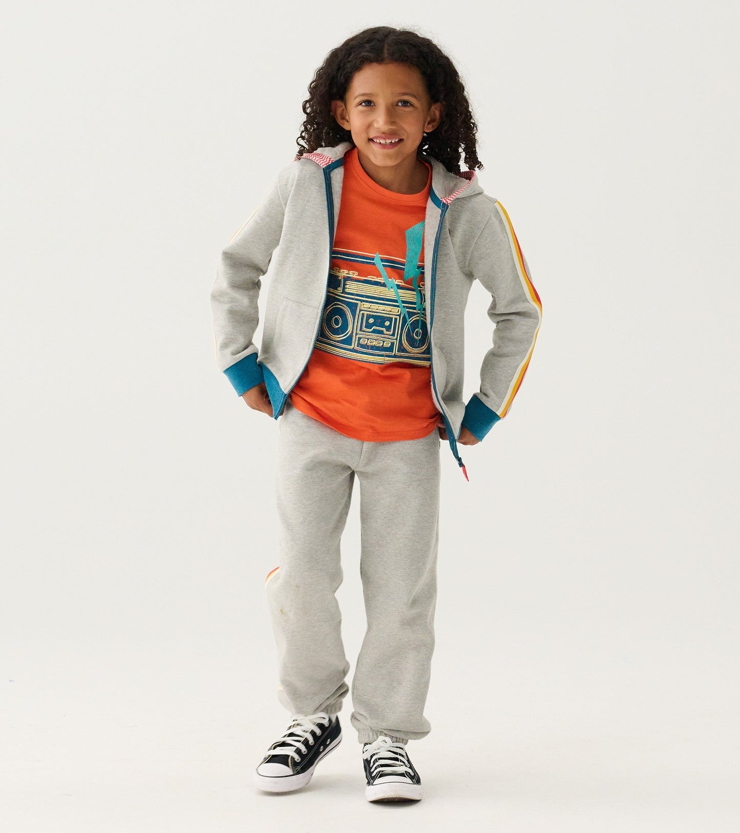 Orange graphic tee featuring a boombox design, paired with gray sweatpants and a gray jacket, worn by a smiling child.