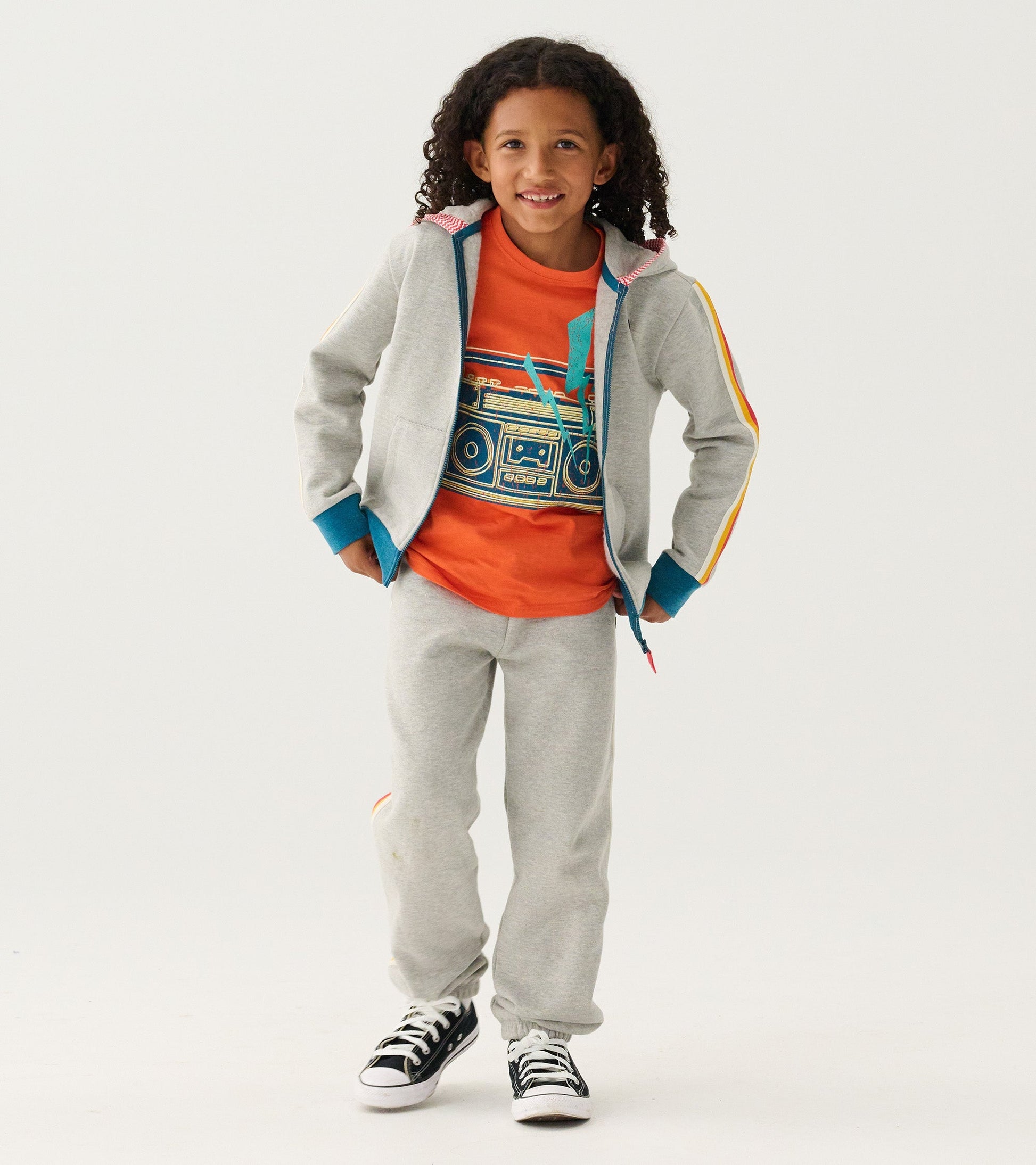 Orange graphic tee featuring a boombox design, paired with gray sweatpants and a gray jacket, worn by a smiling child.