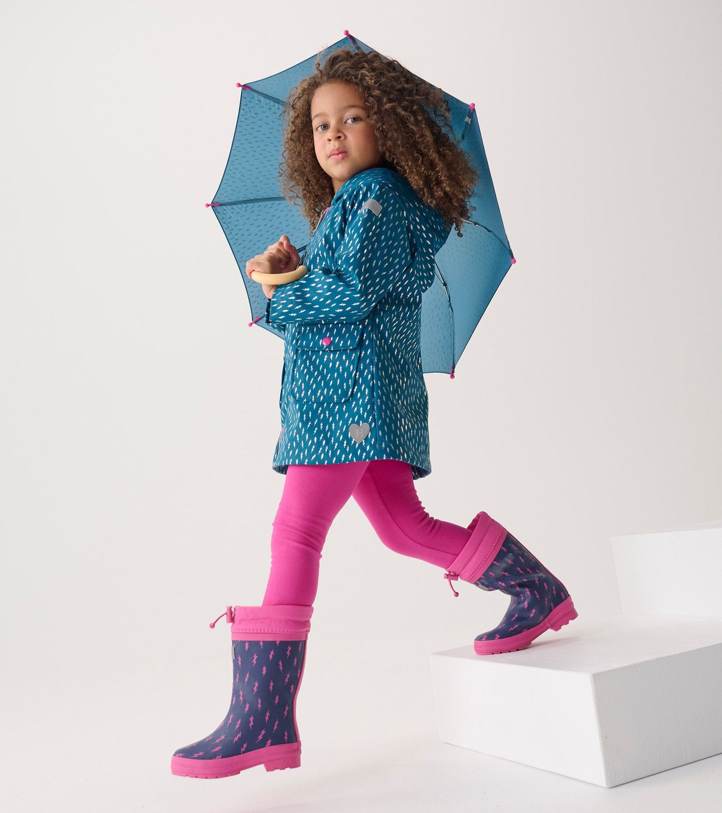 Teal rain jacket with lightning bolt pattern, paired with pink leggings and matching rain boots, under a blue umbrella.