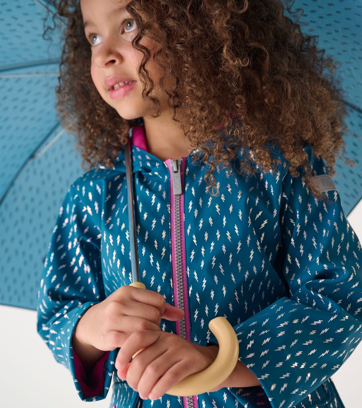 Teal zip-up rain jacket with white lightning bolt pattern, worn by a girl holding a light-colored umbrella.
