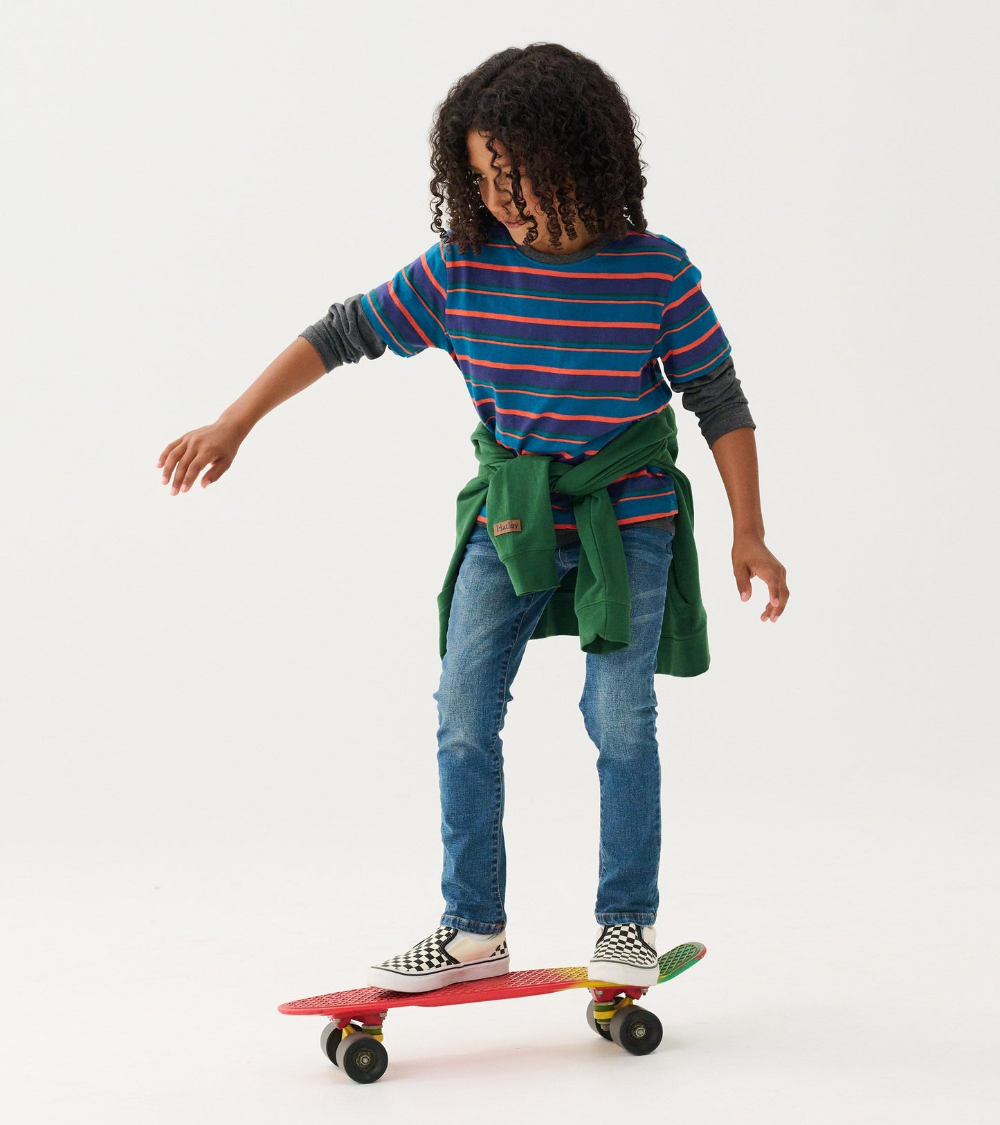 Boys long sleeve t-shirt in blue with colorful stripes, paired with jeans and a green sweatshirt tied around the waist.