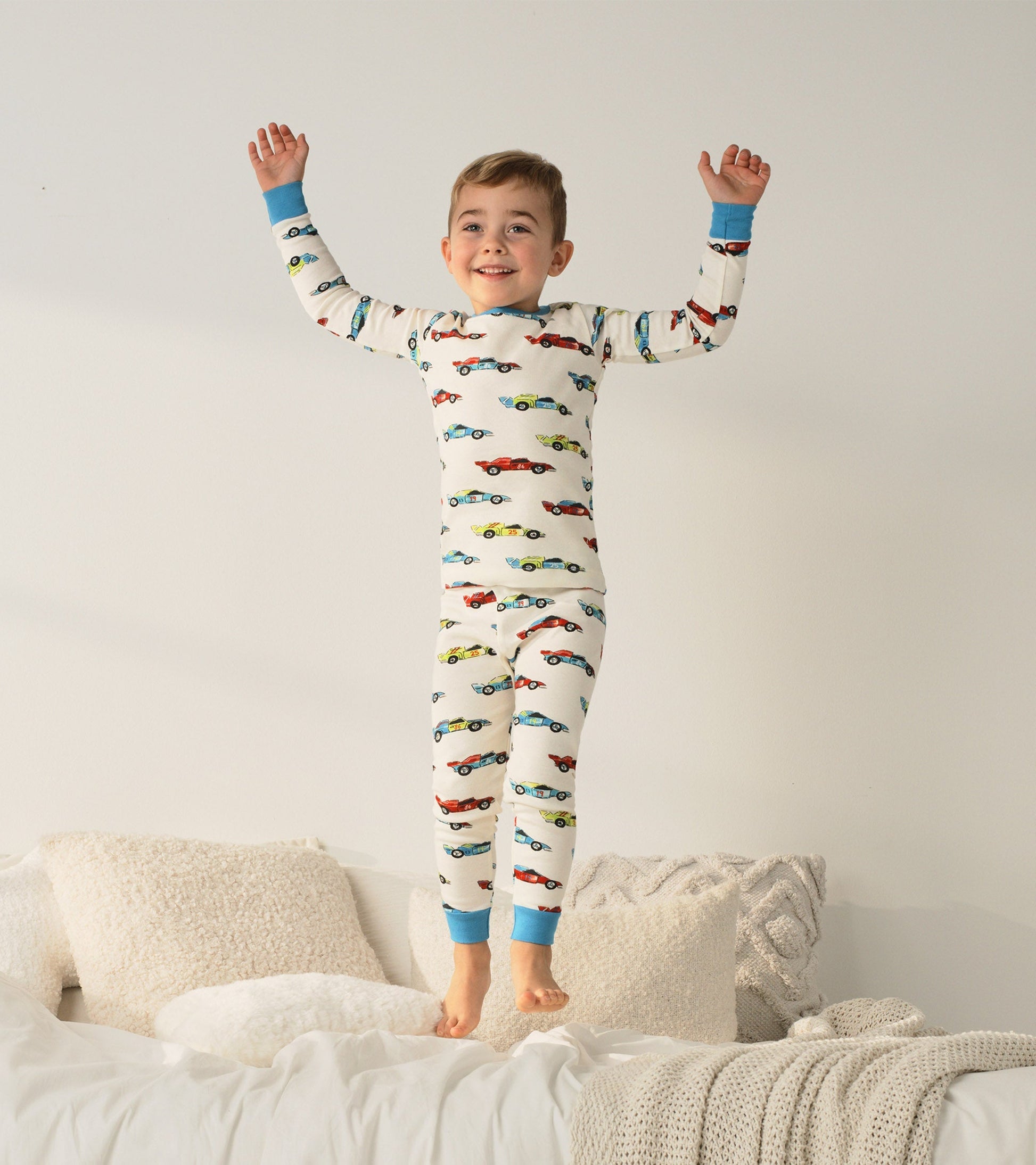 Organic cotton pajama set featuring colorful racecar prints, with blue cuffs, worn by a smiling boy jumping on a bed.