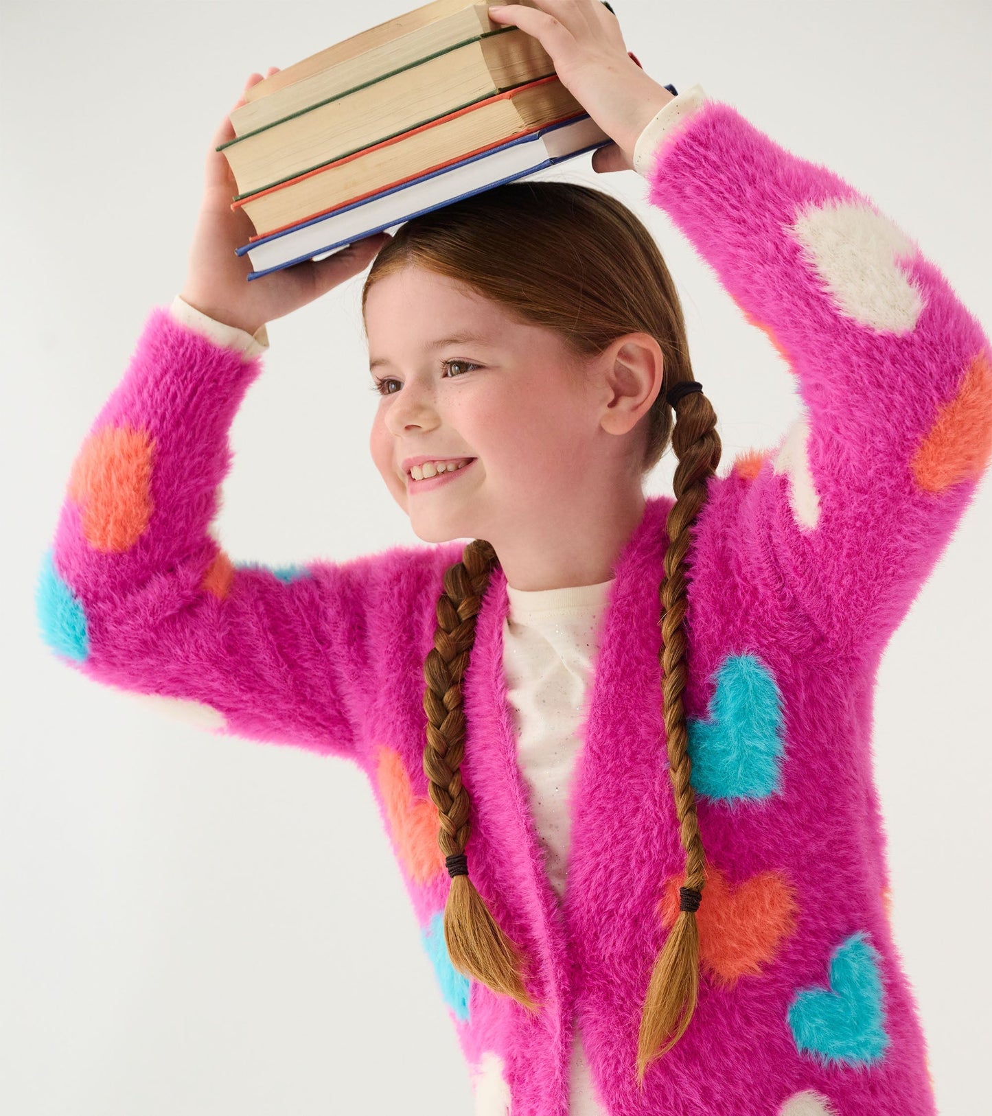 Bright pink cardigan with fluffy texture, featuring colorful heart patterns, worn by a girl holding books above her head.
