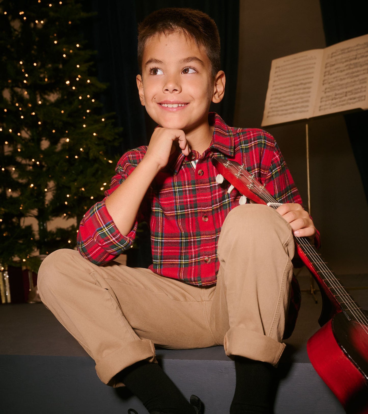 Red plaid button-down shirt on a boy, paired with khaki pants, sitting with a guitar and a festive backdrop.