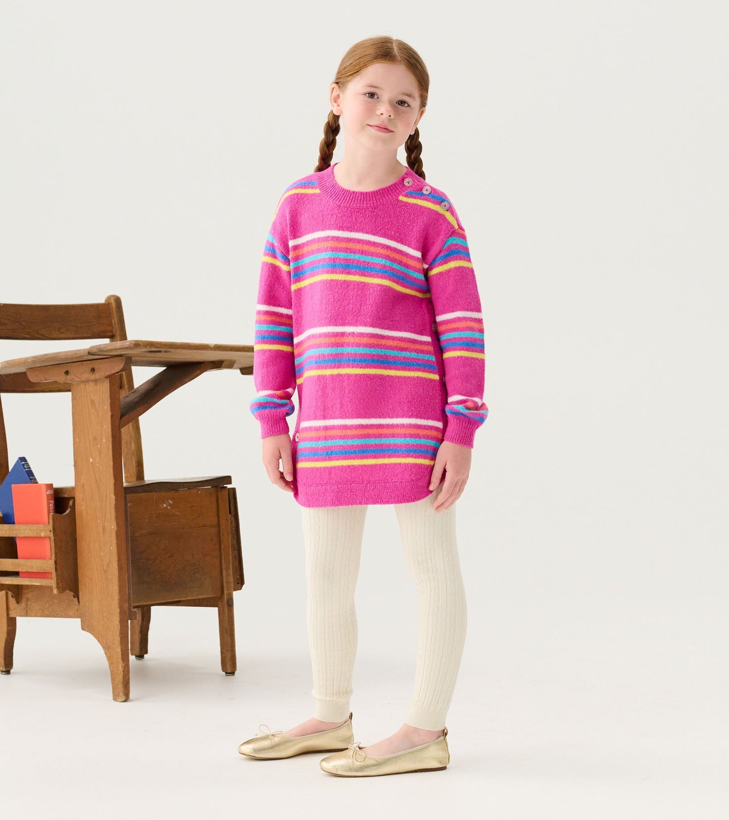 Pink sweater tunic with rainbow stripes, long sleeves, and button detail, paired with cream ribbed leggings and gold flats.