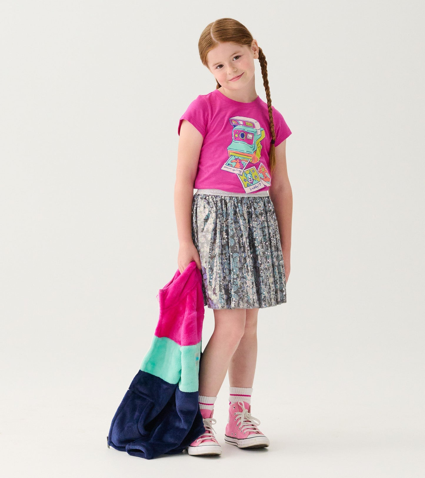 Pink short-sleeve tee with a colorful graphic, paired with a shiny silver skirt and holding a multicolored jacket.