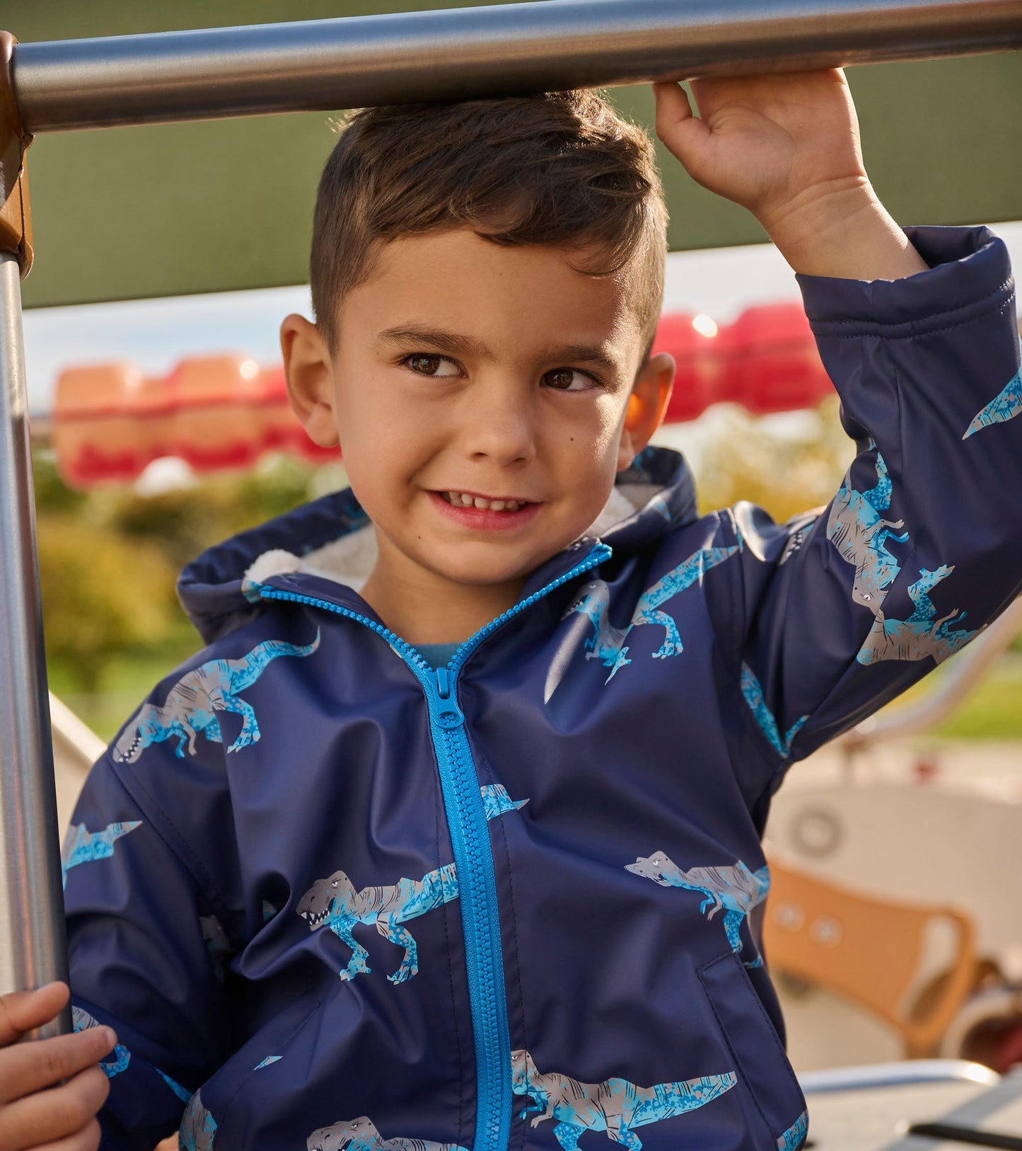 Navy rain jacket with T-Rex print, zip-up front, and hood, worn by a smiling boy on a playground.