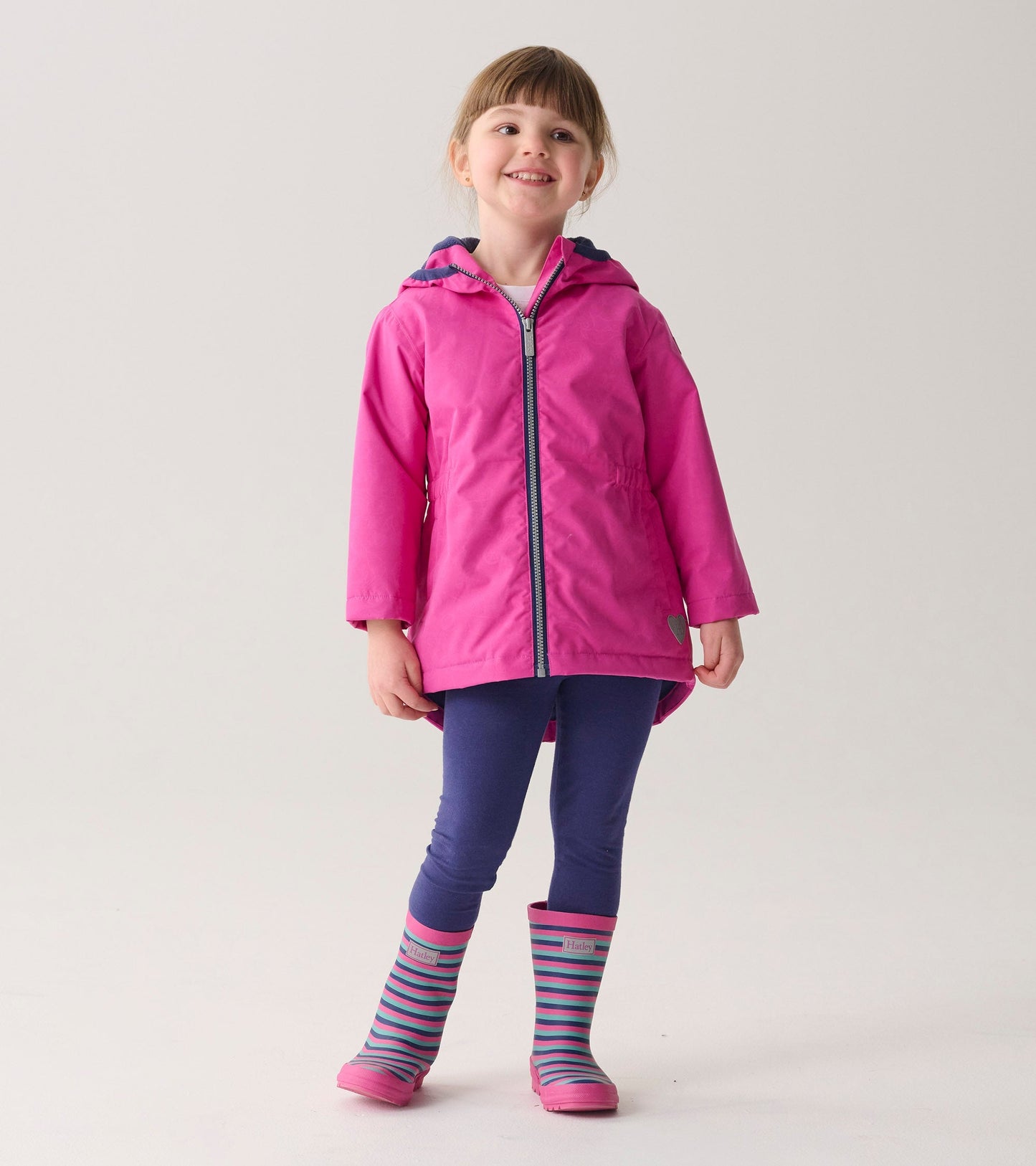 Lightweight pink zip-up rain jacket with a hood, worn by a girl, paired with navy leggings and striped rain boots.