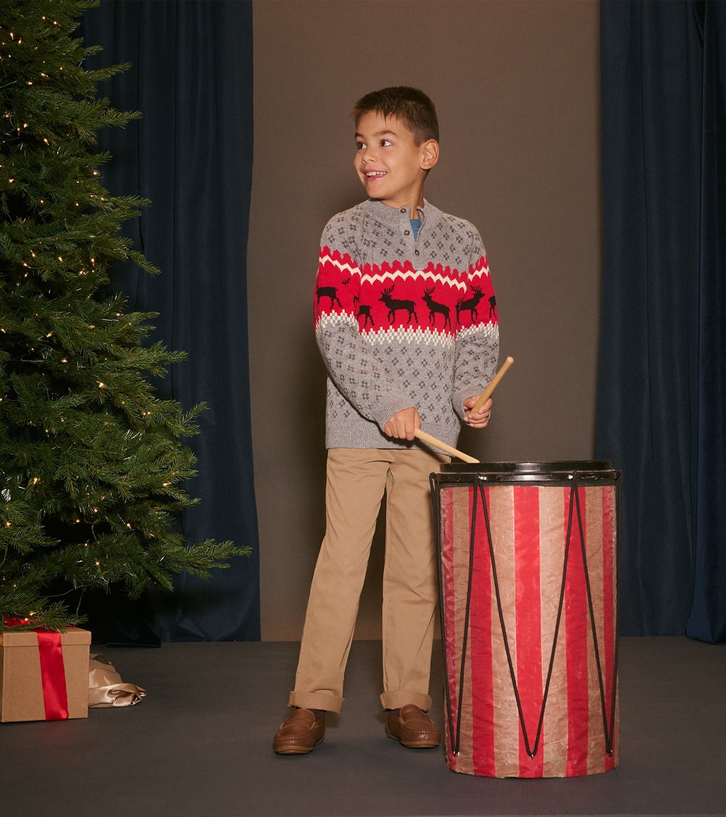Gray mockneck sweater with red elk pattern, worn by a boy playing a drum, festive setting with a Christmas tree.