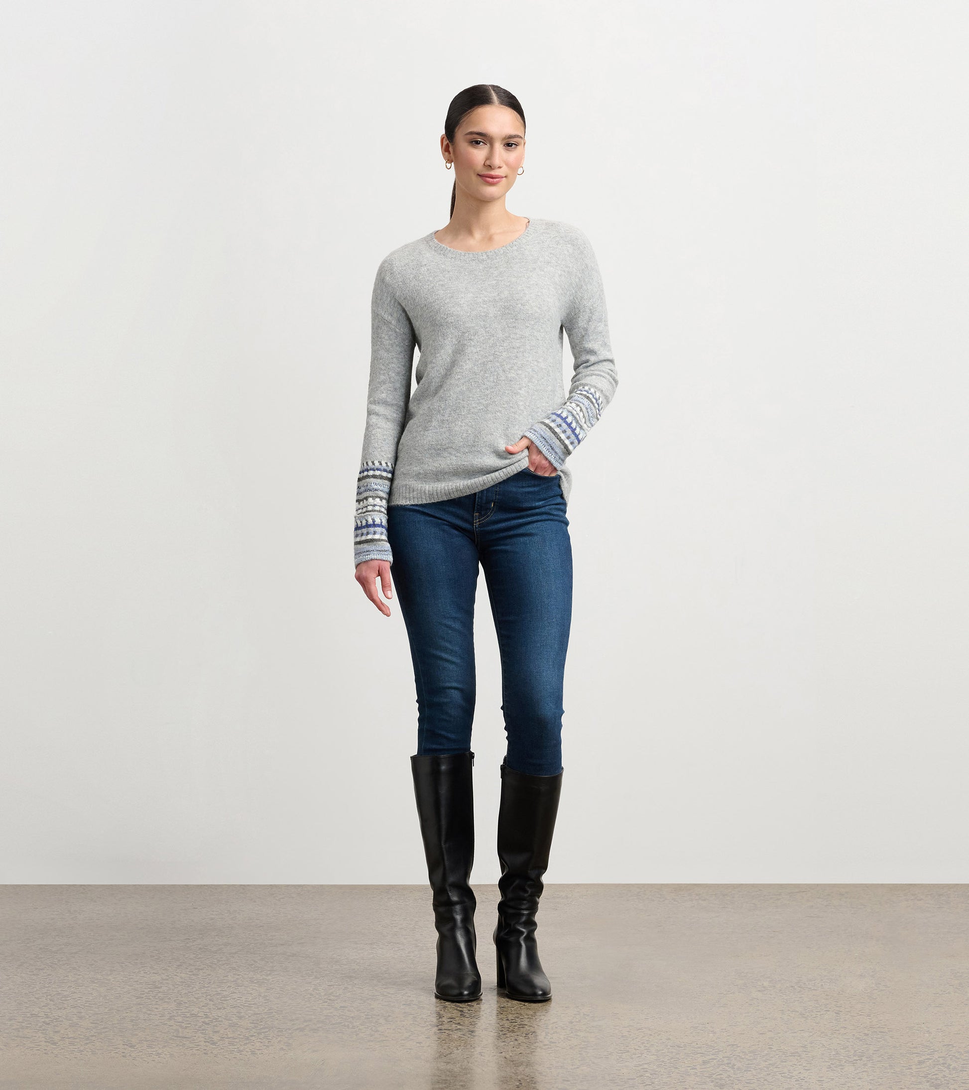Grey melange sweater with patterned cuffs, featuring a relaxed fit and round neckline, paired with dark jeans and knee-high…