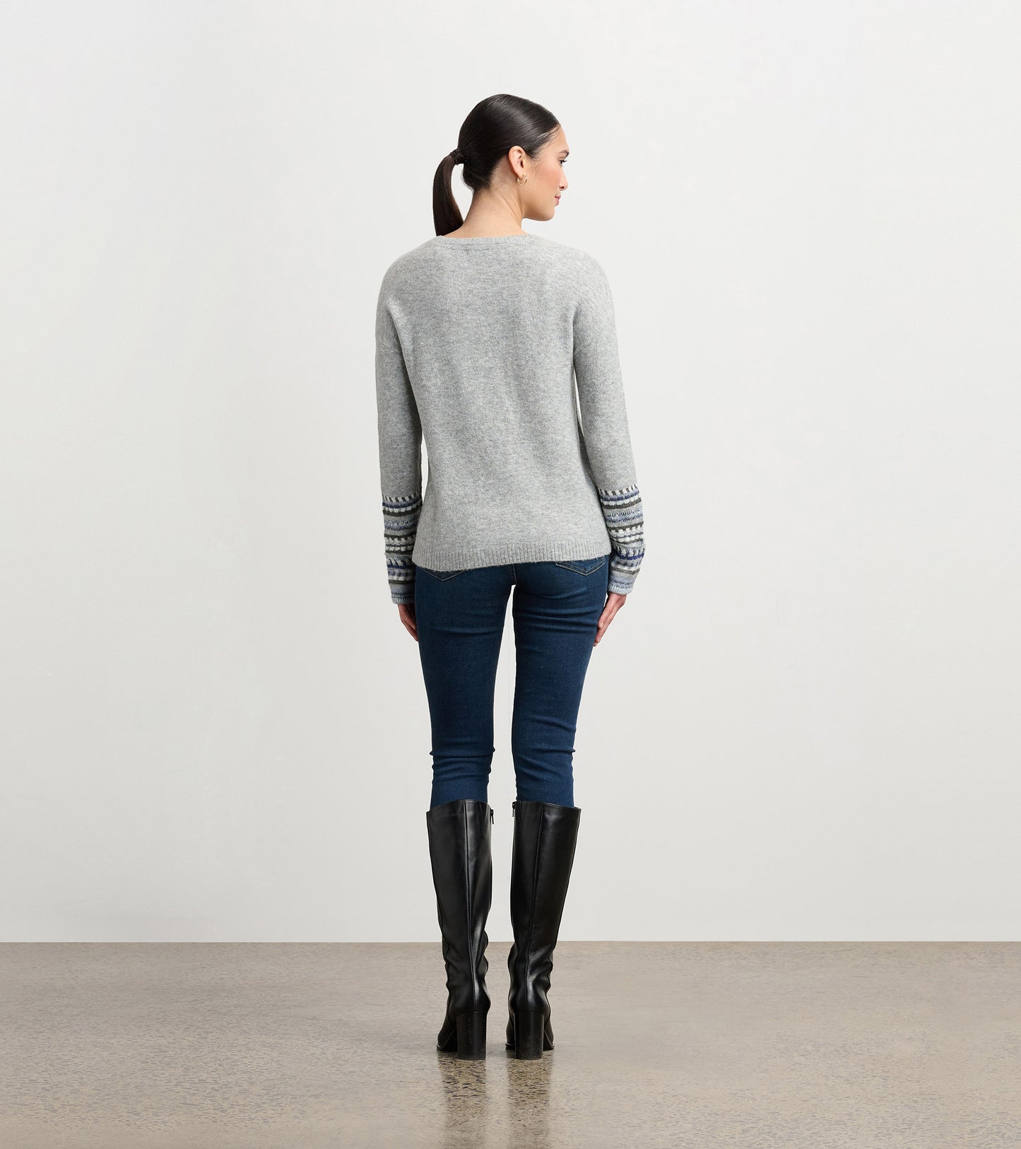 Grey melange sweater with jacquard cuffs, long sleeves, and a relaxed fit, worn with dark jeans and knee-high boots.
