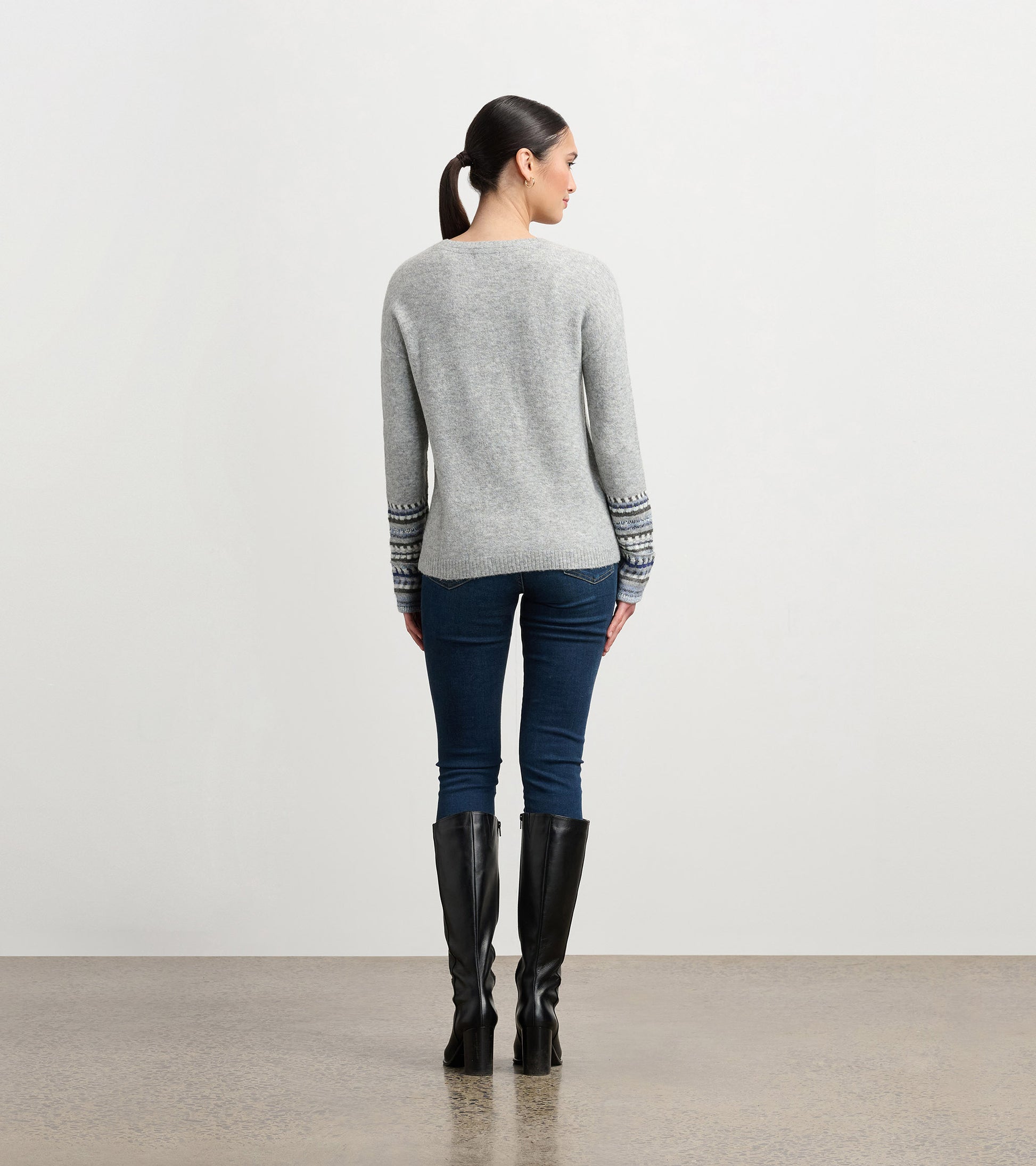 Grey melange sweater with jacquard cuffs, long sleeves, and a relaxed fit, worn with dark jeans and knee-high boots.
