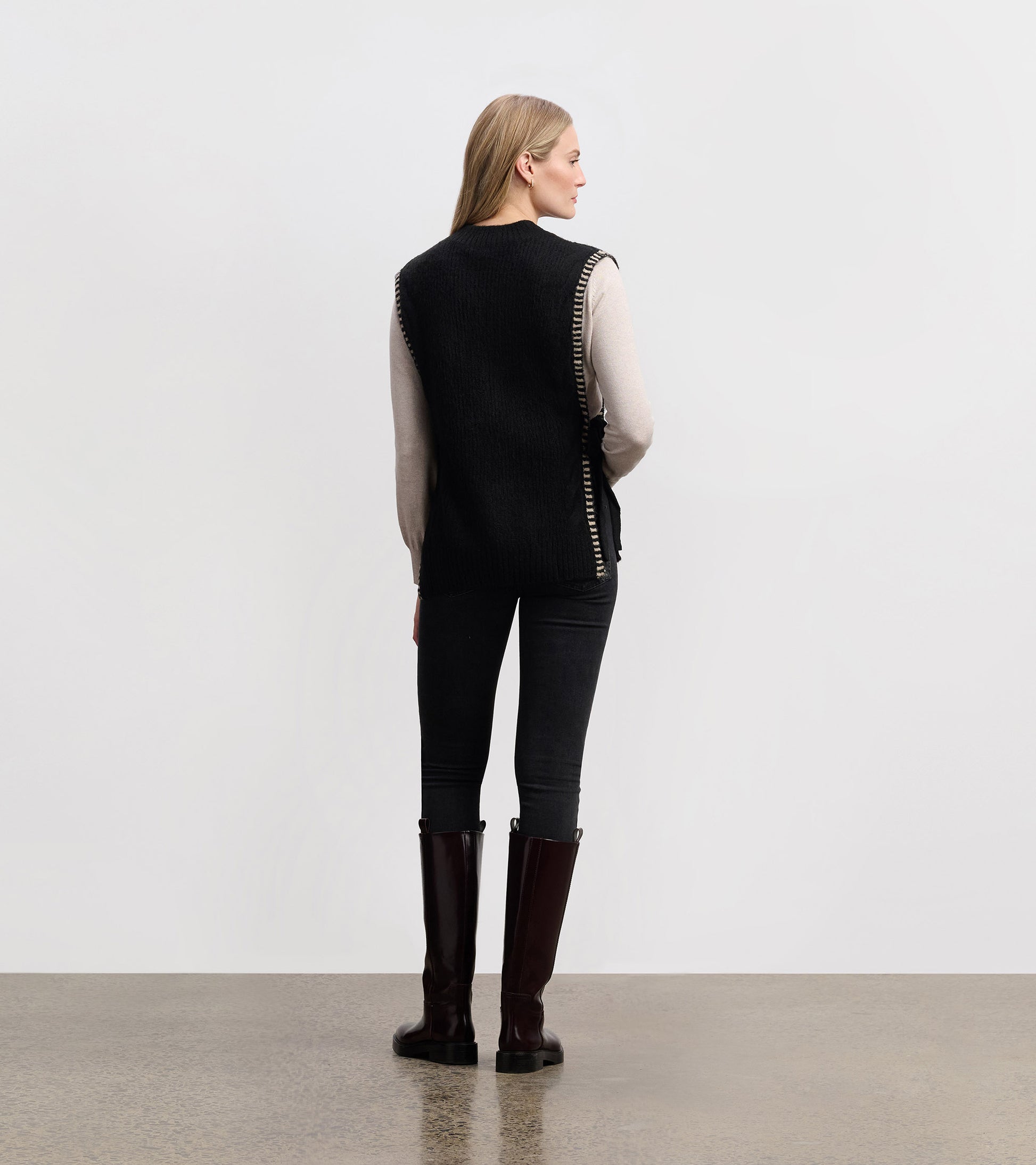 Black side-tie sweater vest with a textured knit, worn over a light top, styled with black jeans and tall boots.