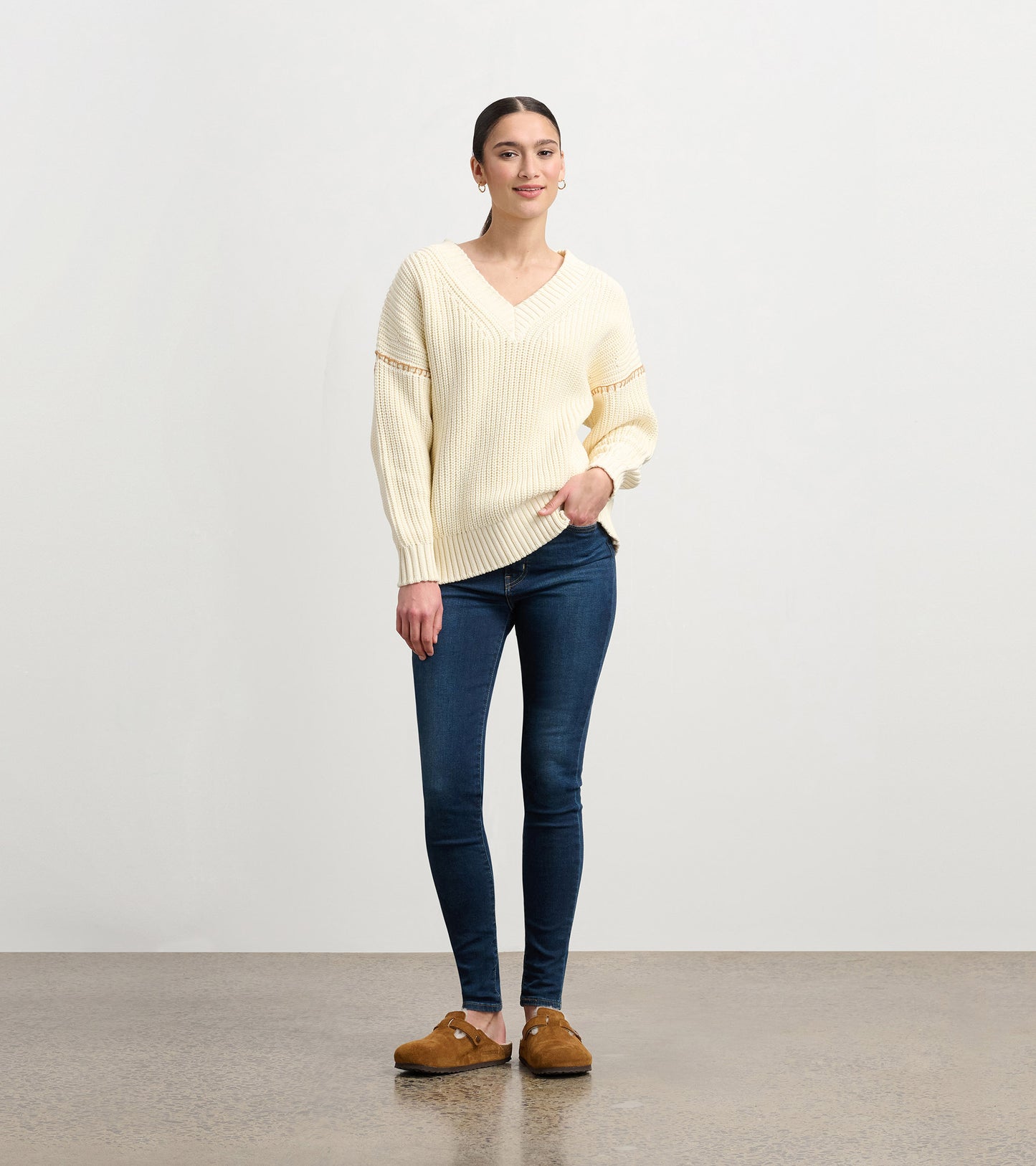 Cream V-neck slouchy sweater with ribbed texture, paired with dark jeans and brown clogs, styled casually.