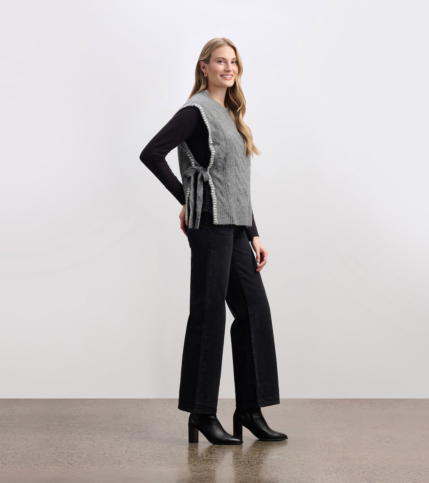 Charcoal melange side-tie sweater vest with a textured knit, styled over a black long-sleeve top and paired with wide-leg pa…