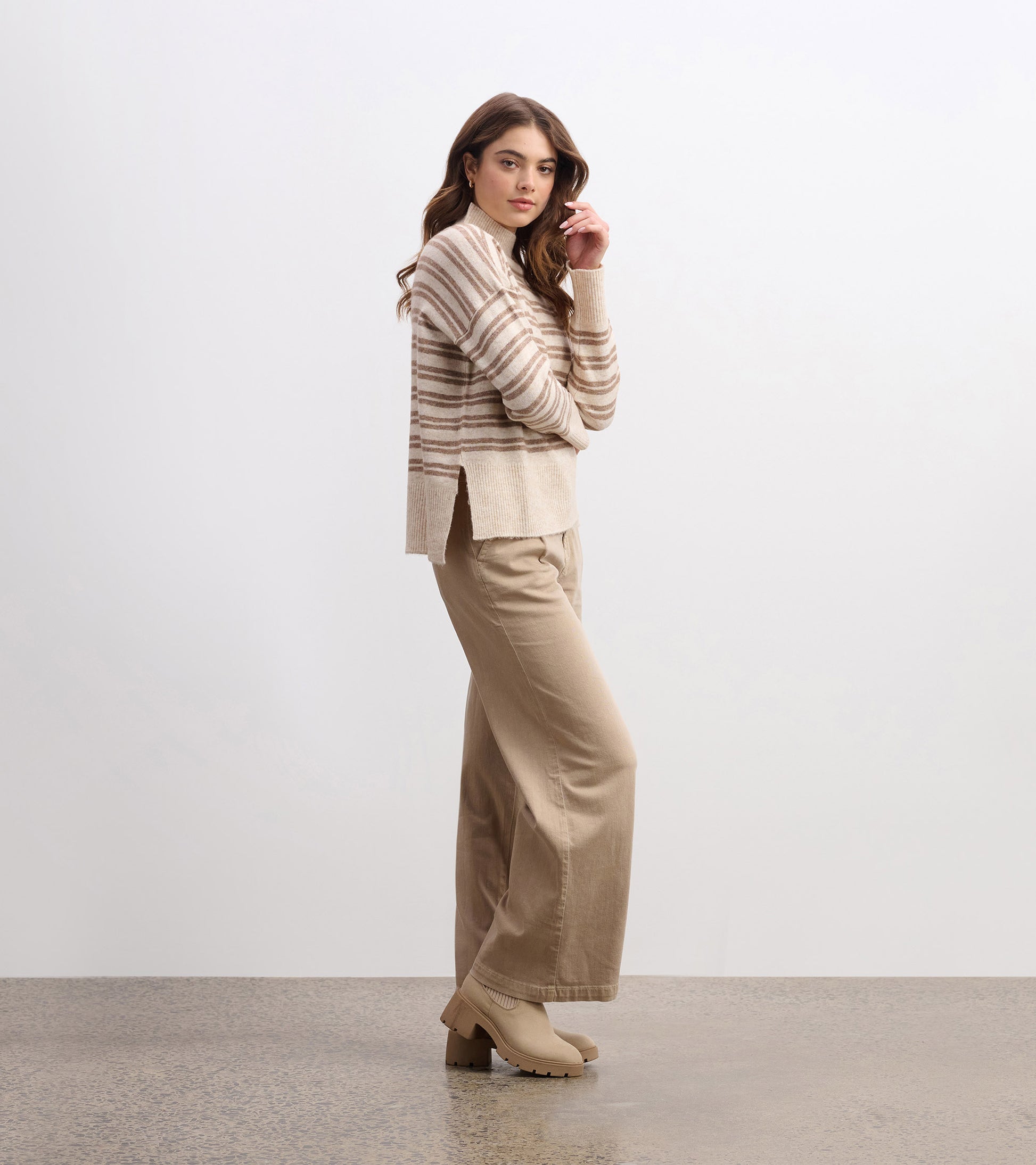 Mock neck sweater in camel with double stripes, featuring a relaxed fit and side slits, styled with beige wide-leg pants.