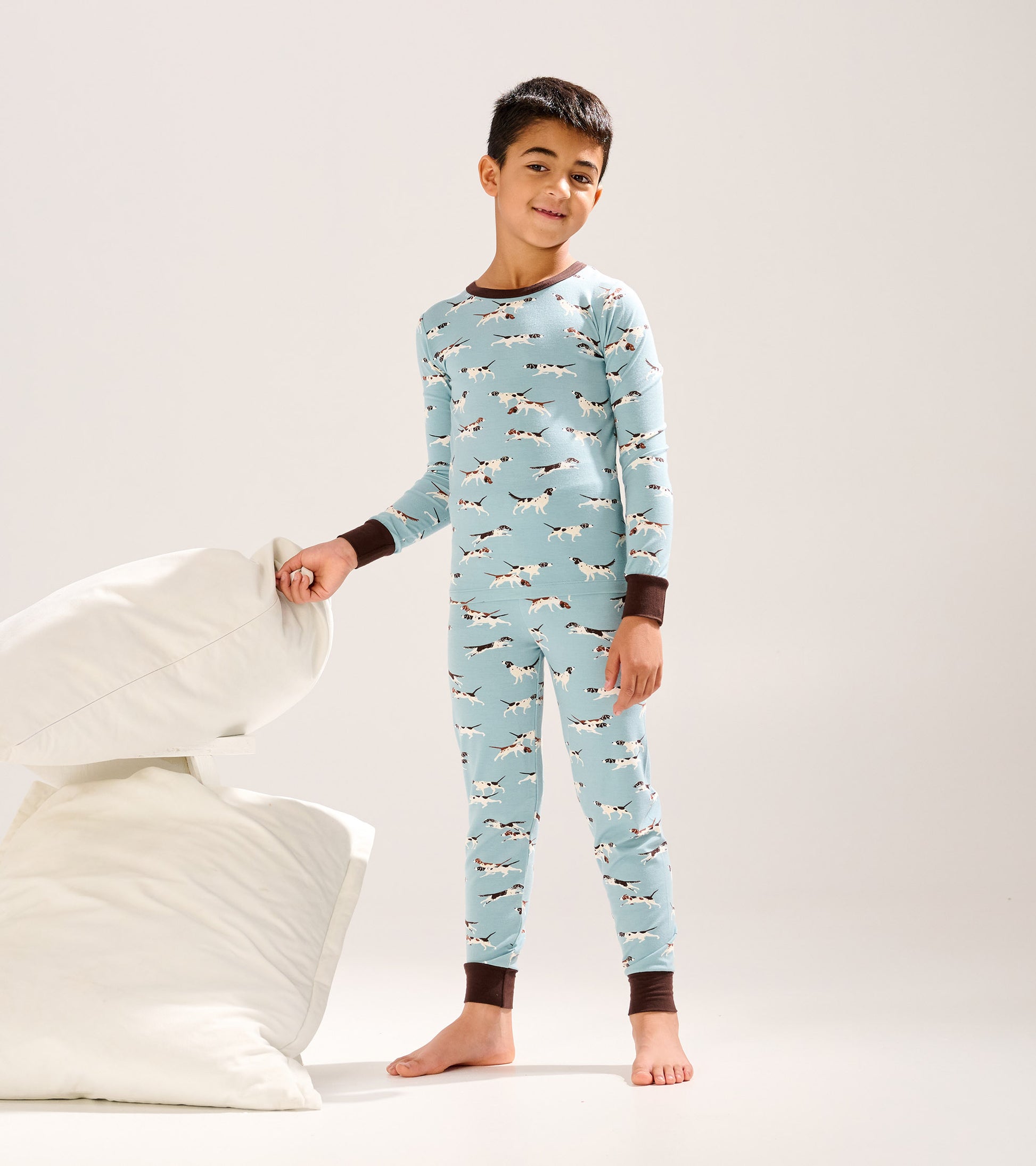 Light blue bamboo pajama set featuring hunting dogs print, with brown cuffs, worn by a boy standing beside pillows.