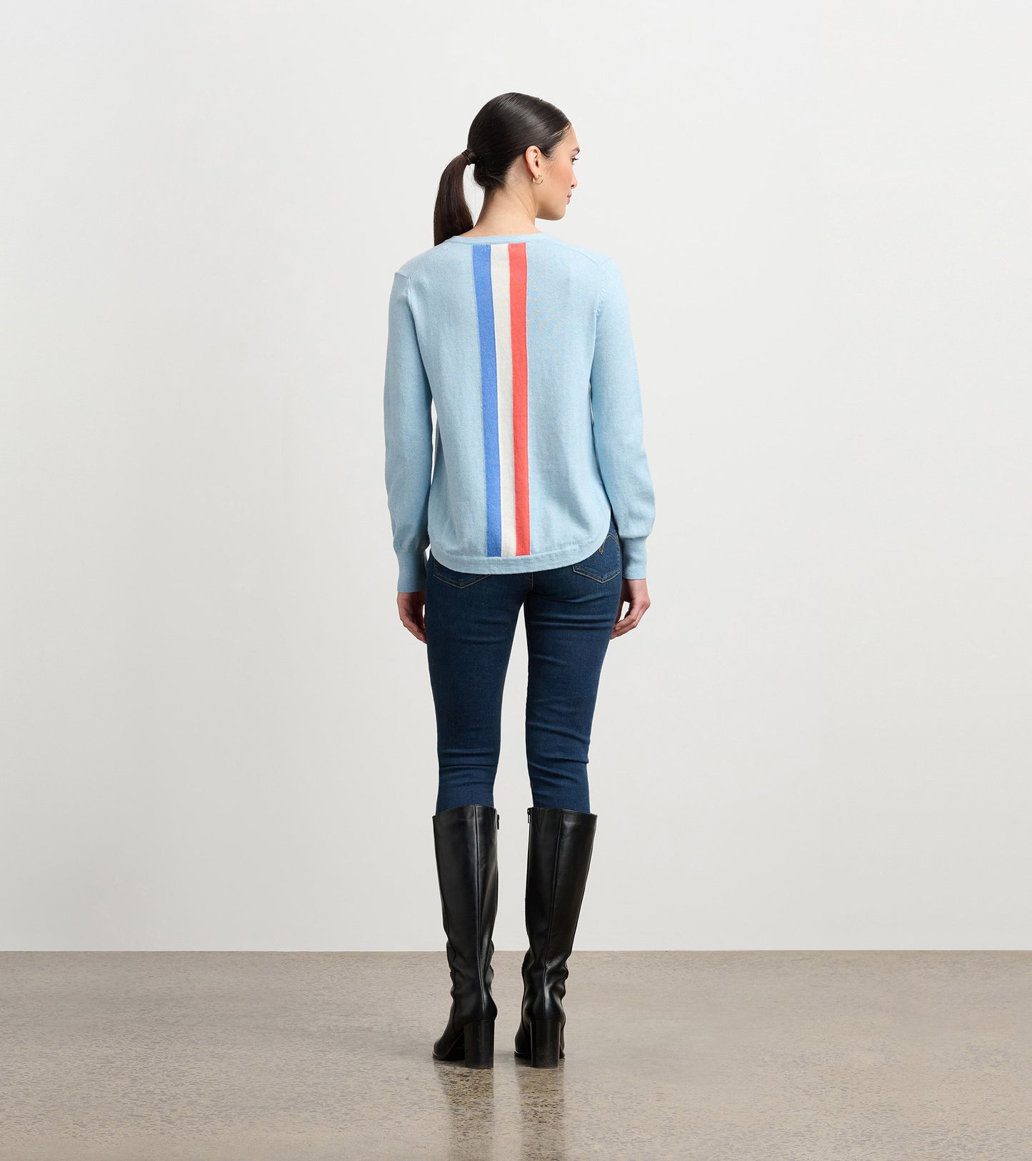 Light blue sweater with a striped back design in red and blue, paired with dark jeans and knee-high black boots.