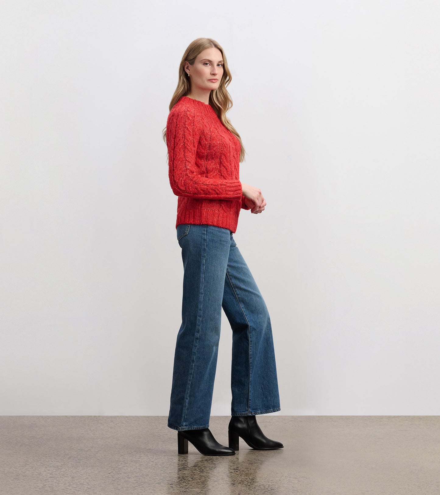 Red cable knit sweater with long sleeves, styled with high-waisted jeans and black heeled boots, shown from the side.