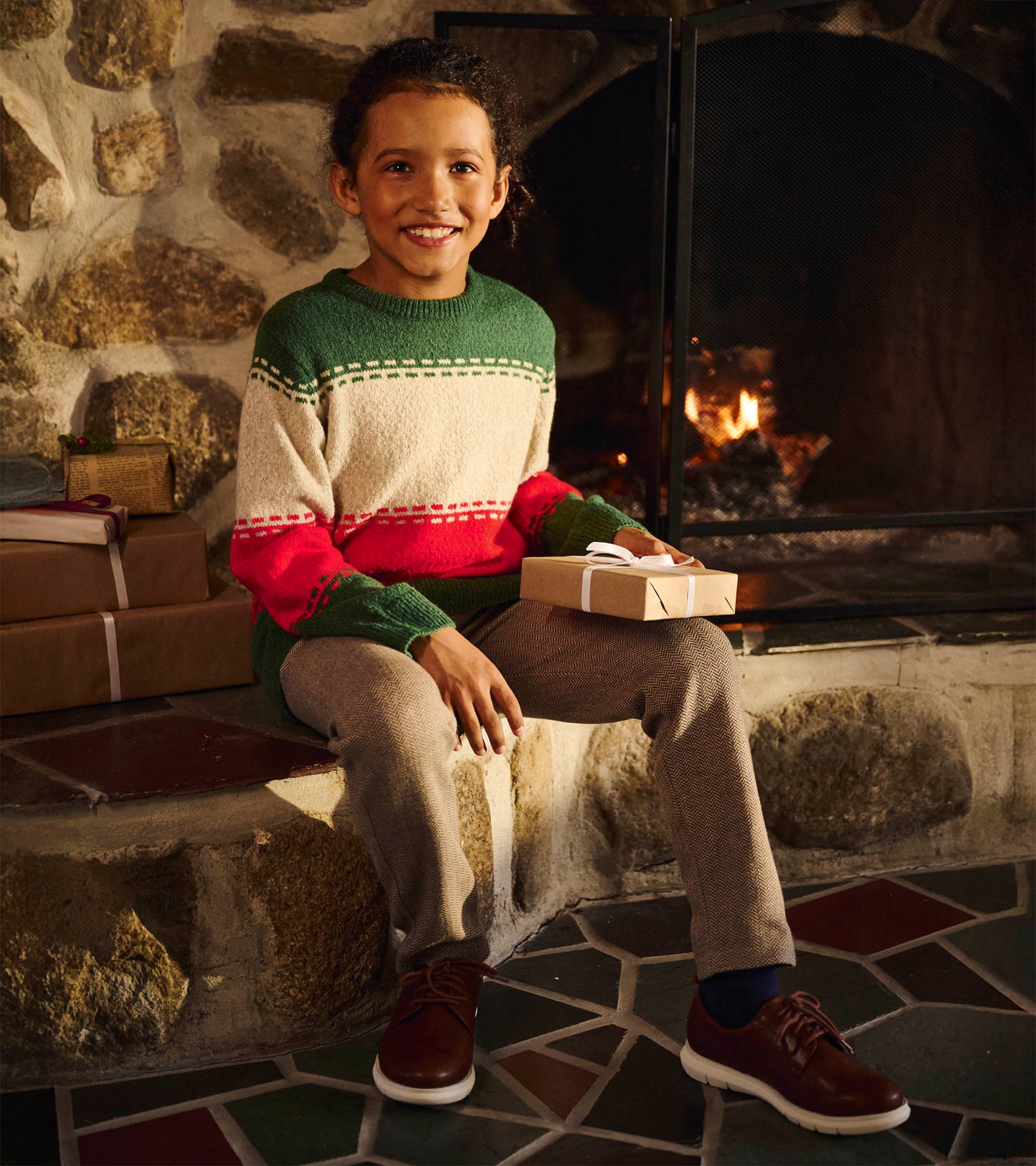 Boy wearing a green, cream, and red striped crew neck sweater with holiday pattern, seated by a fireplace