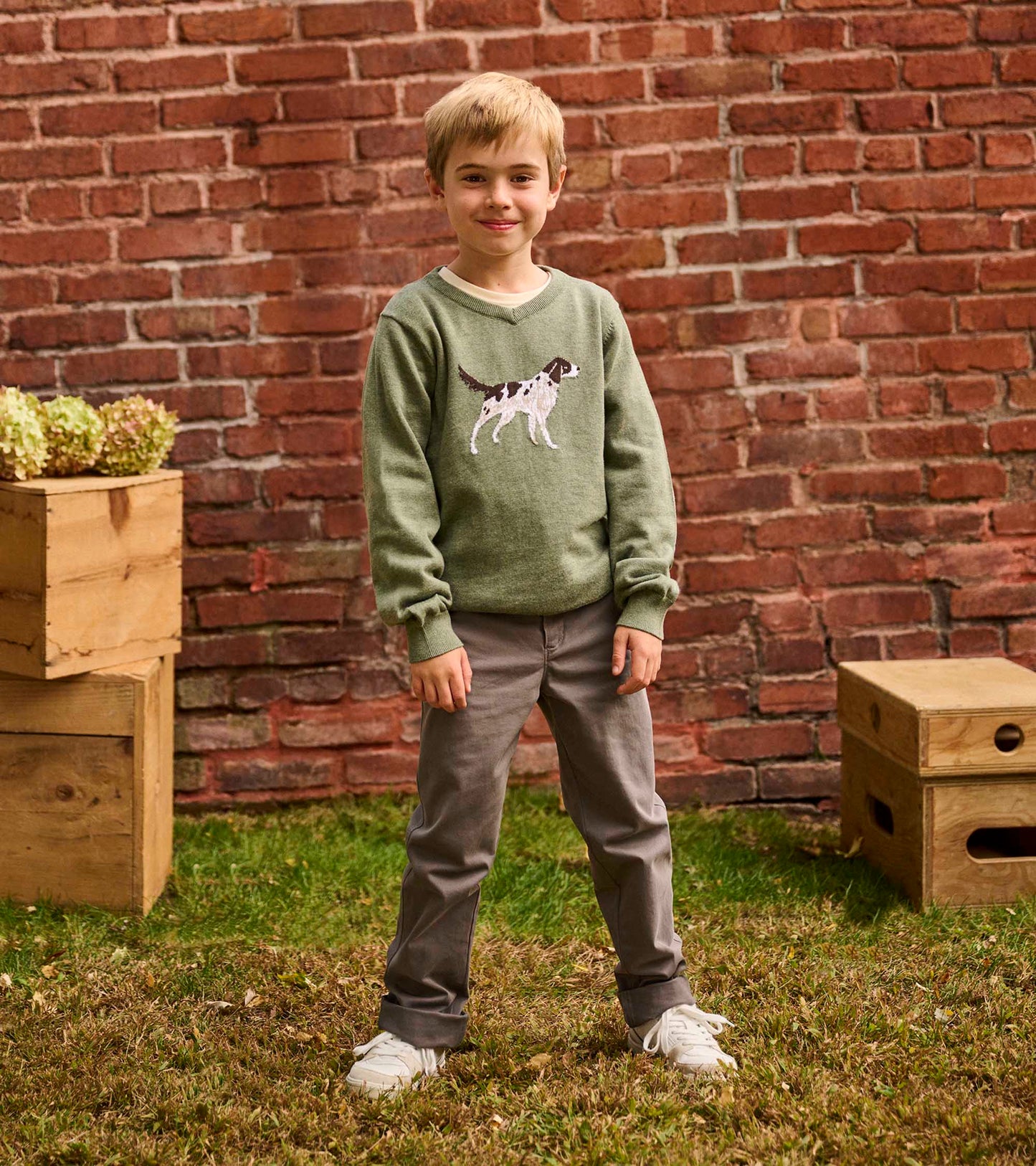 Green V-neck sweater featuring an embroidered hunting dog, worn by a boy with gray pants and white sneakers.