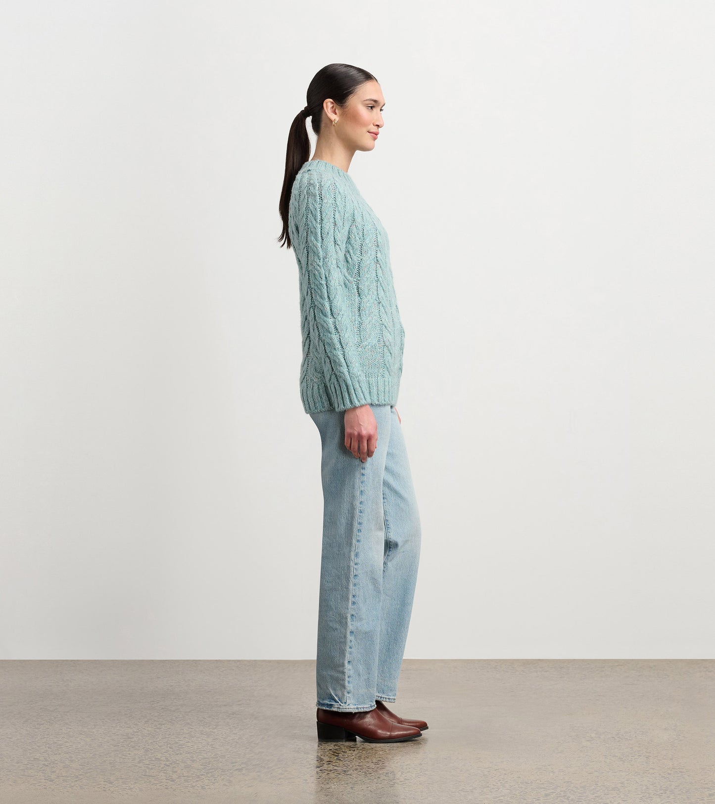 Light turquoise cable knit sweater with a relaxed fit, worn with light blue jeans and brown boots, viewed from the side.