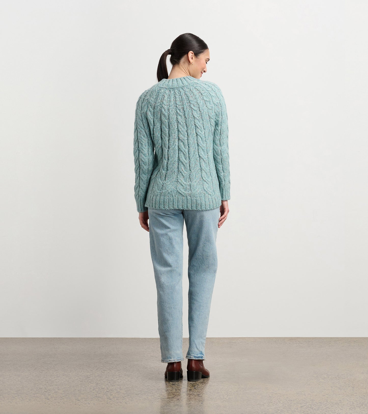 Back view of a light turquoise cable knit sweater with ribbed trim and long sleeves, worn with jeans.