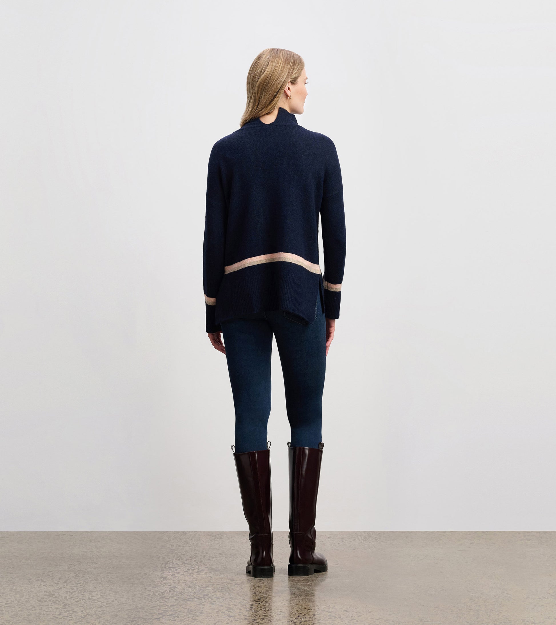 Navy mock neck sweater with a striped hem, featuring a relaxed fit and side slits, worn with dark jeans and tall boots.