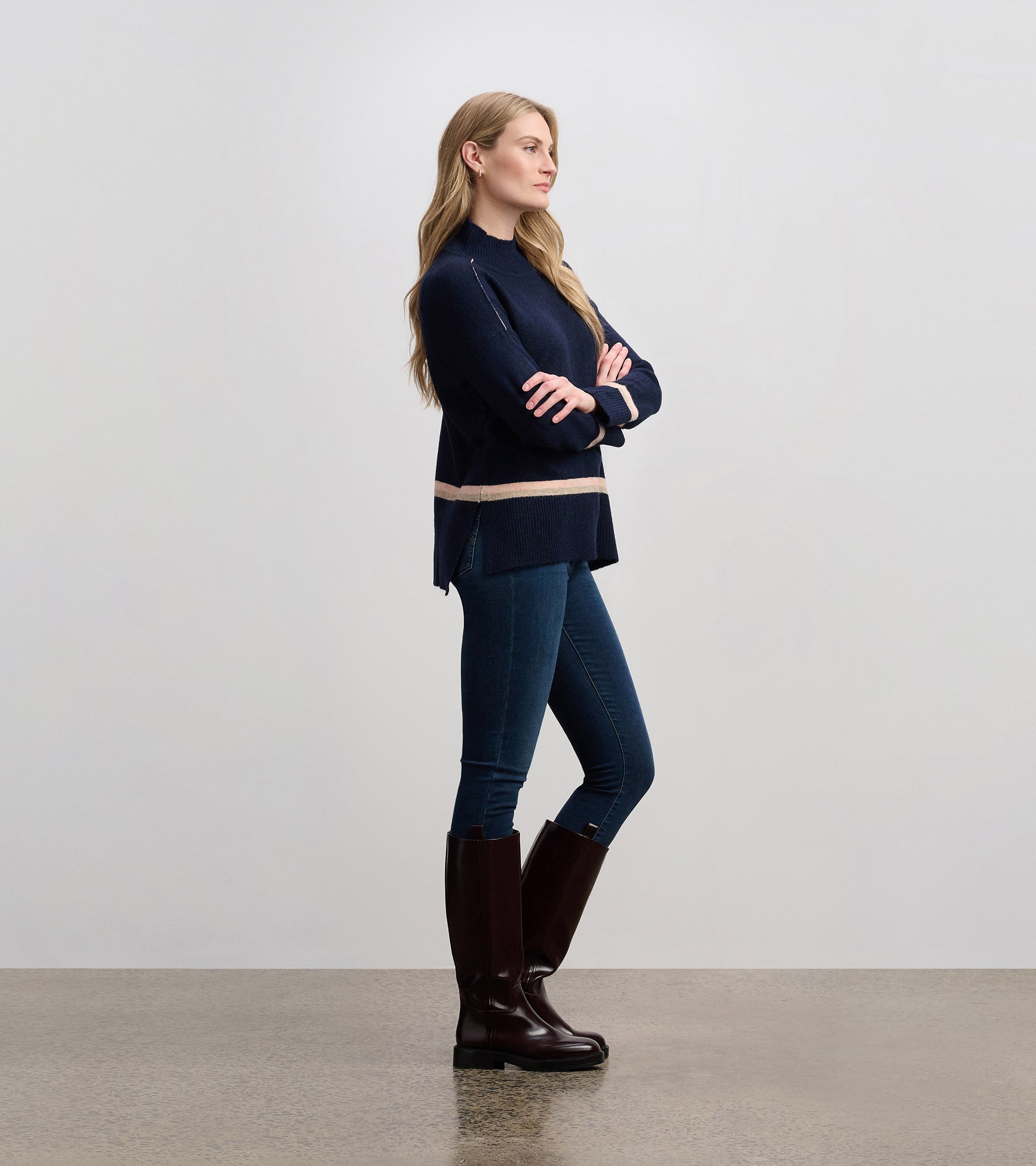 Navy mock neck sweater with a striped hem, paired with fitted jeans and knee-high boots, shown from the side.