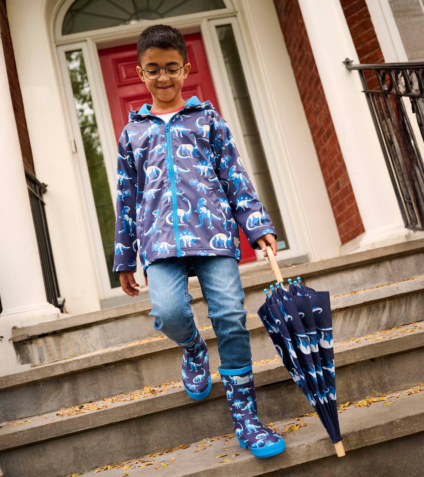 Boys' zip-up rain jacket featuring a blue dinosaur pattern on a dark background, paired with blue rain boots and an umbrella.