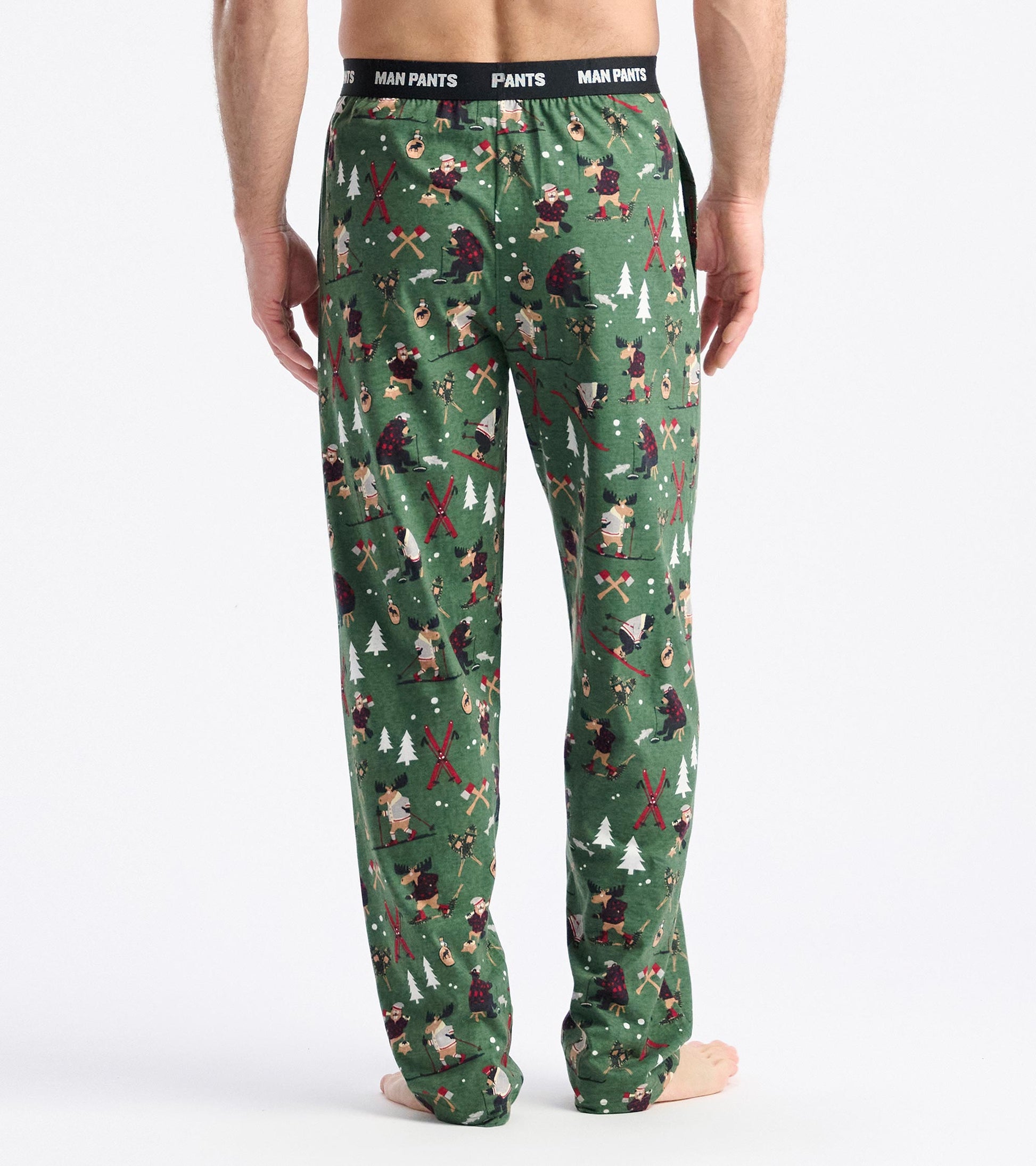 Green pajama pants featuring a playful winter-themed pattern with trees, animals, and outdoor activities, viewed from the ba…