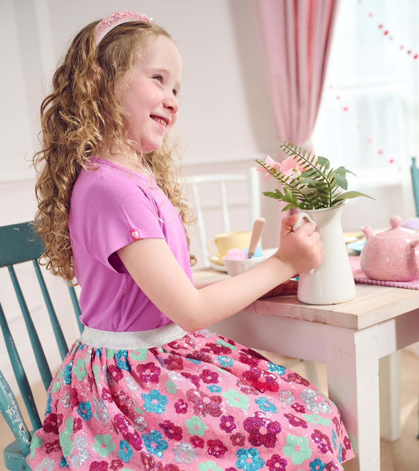 Purple short-sleeve tee worn by a girl, paired with a colorful floral skirt, sitting at a table with a vase.
