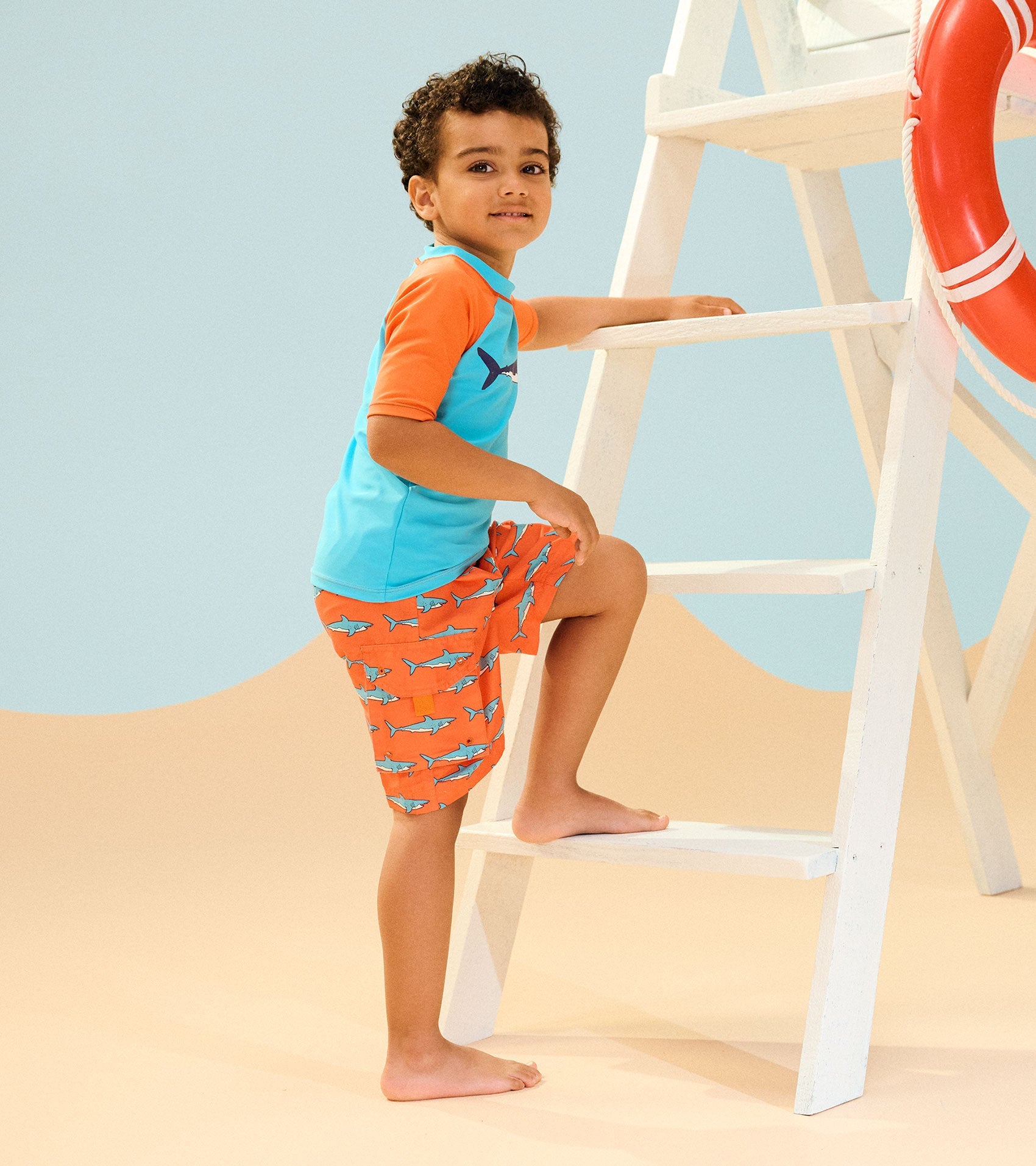 Short sleeve rashguard in blue and orange with shark pattern, paired with matching shorts, worn by a boy on a ladder.