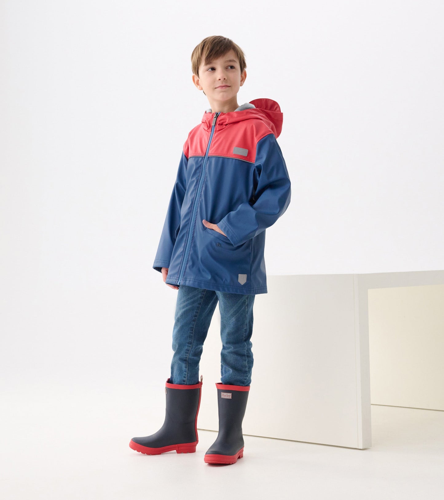 Red and navy zip-up rain jacket with a hood, featuring pockets and a relaxed fit, worn by a boy with jeans and rain boots.