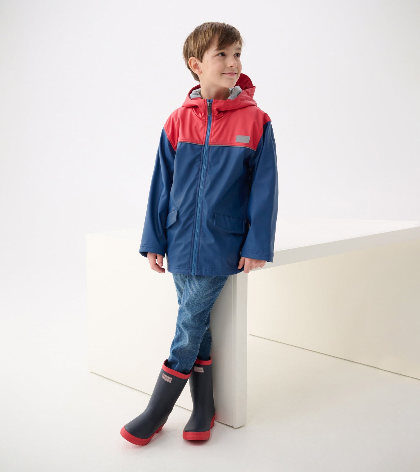 Boys' zip-up rain jacket in red and navy with a hood, featuring two front pockets, worn with jeans and rain boots.