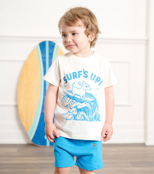 Cream graphic tee featuring a blue surf design, paired with bright blue shorts, worn by a toddler.