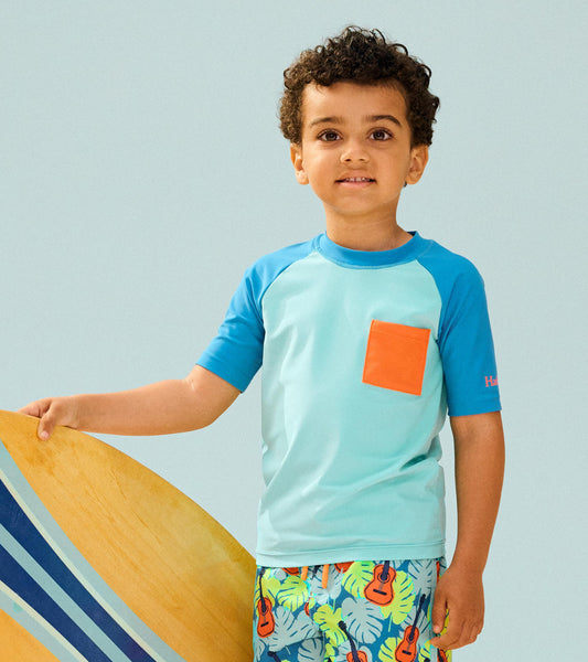 Light blue short sleeve rashguard with a bright orange pocket, worn by a boy holding a surfboard.